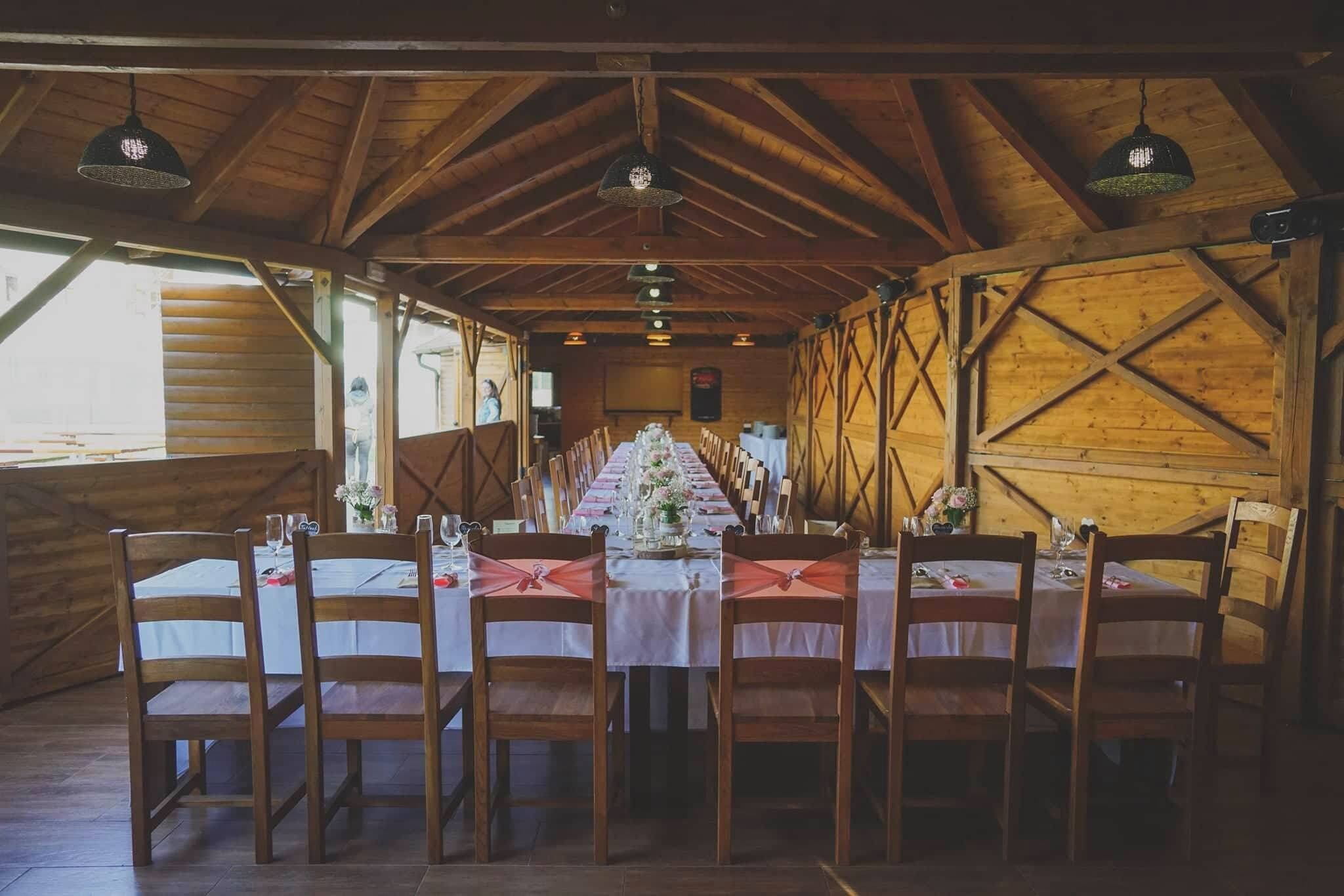 Una larga mesa de comedor de madera dispuesta para una comida, con sillas, vigas de madera en el techo y lámparas colgantes.