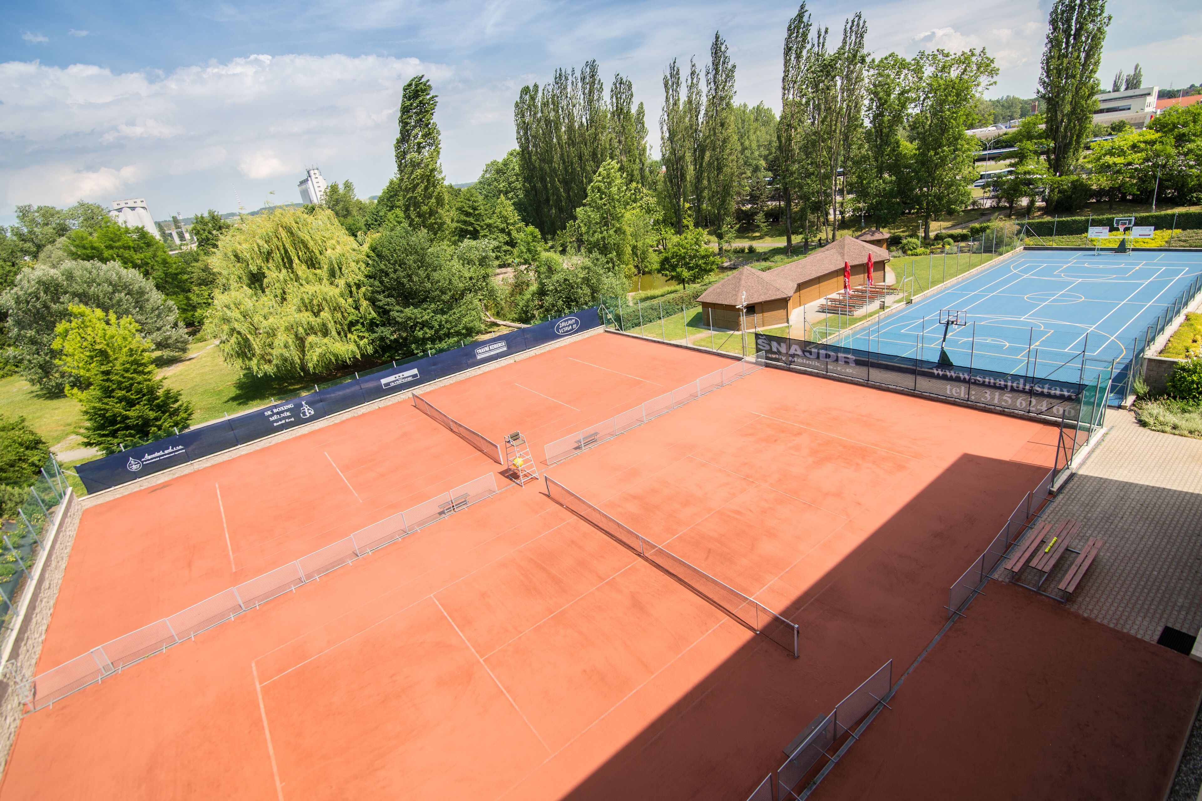 A tennis court with a net, surrounded by trees and a building in the background.