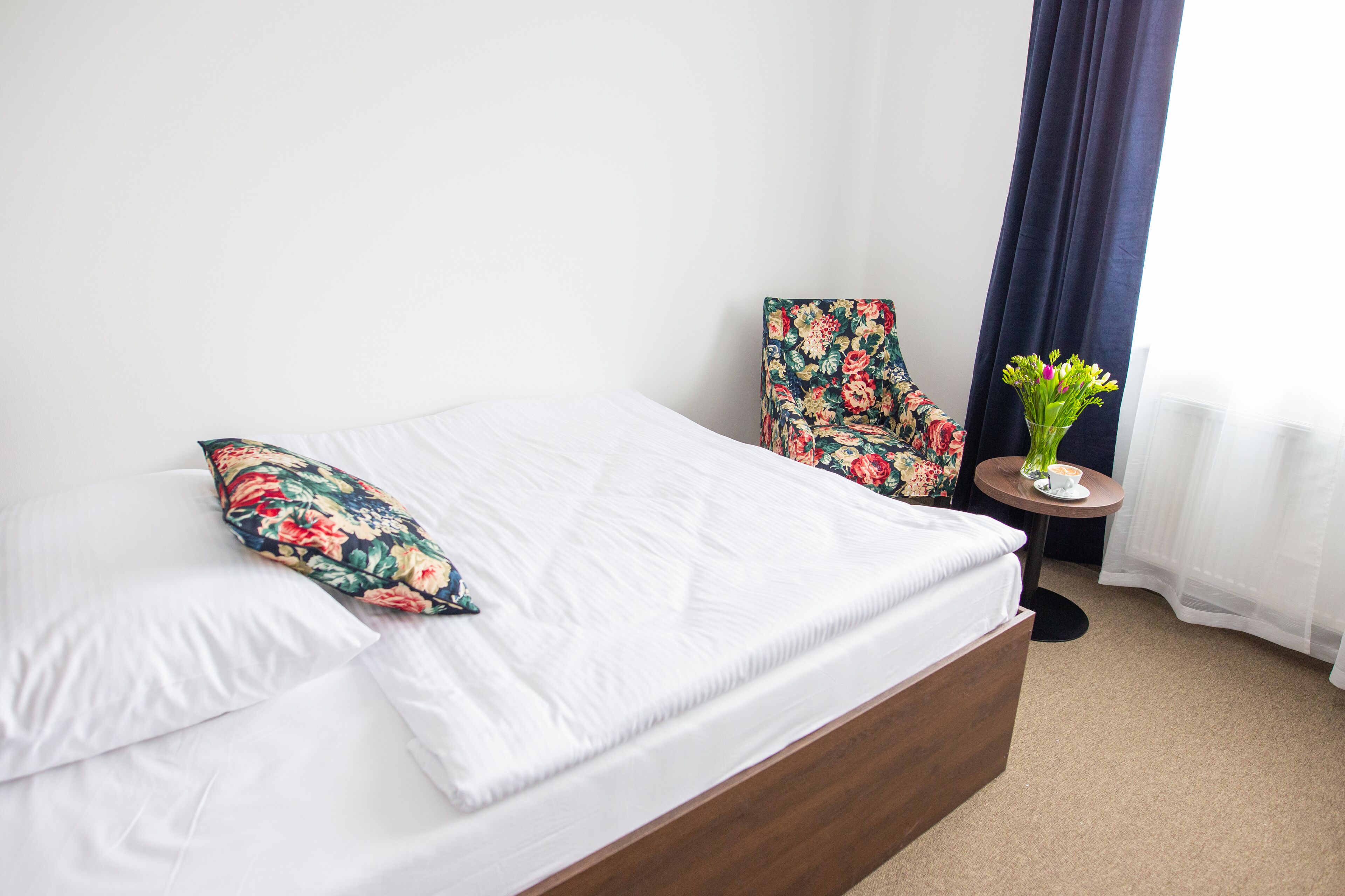 A neatly made bed with white linens and a floral pillow, a floral armchair, a small round table with a vase of flowers, and a dark curtain partially drawn.