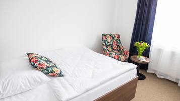 A neatly made bed with white linens and a floral pillow, a floral armchair, a small round table with a vase of flowers, and a dark curtain partially drawn.