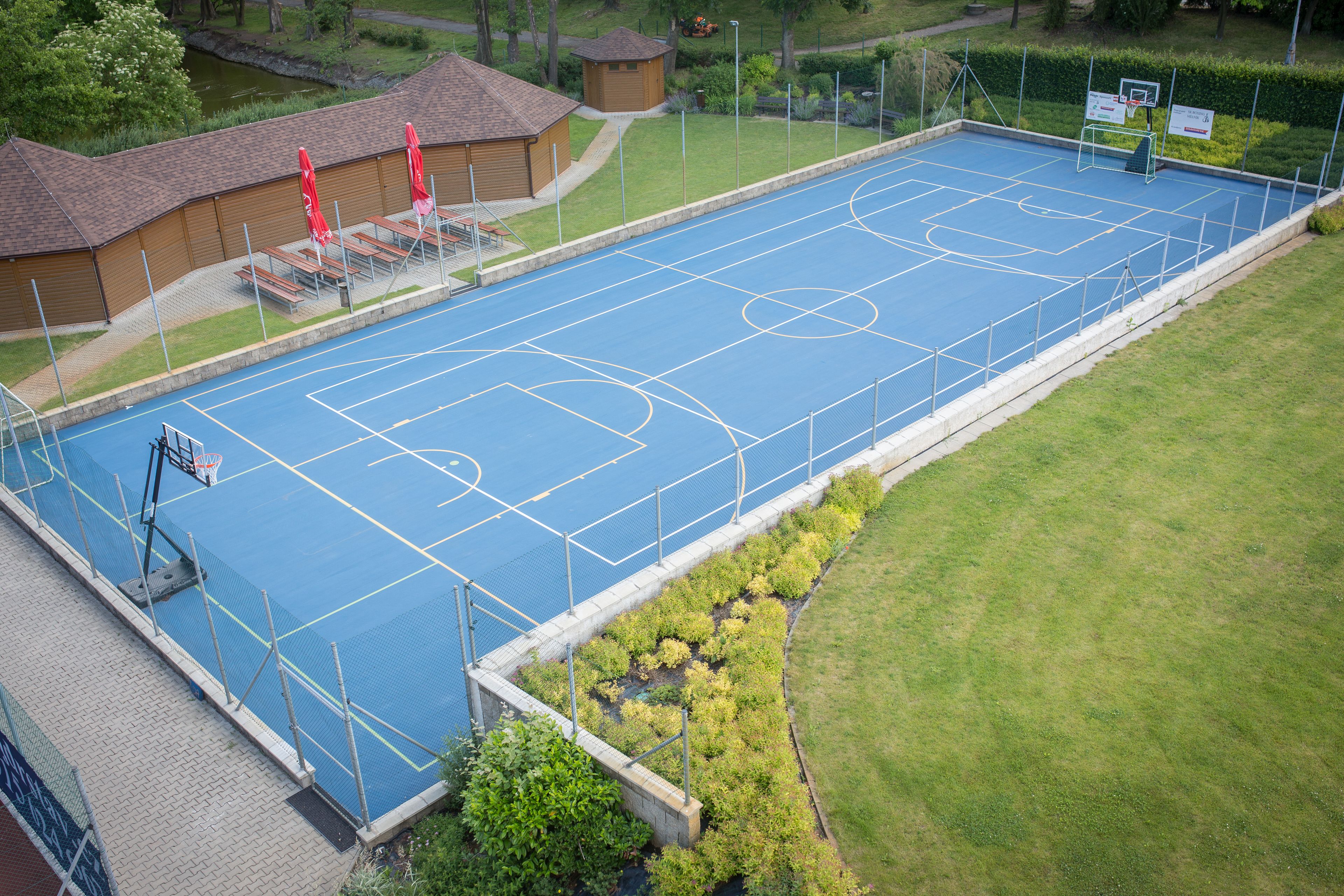 Een buitenbasketbalveld met een blauw oppervlak, omringd door een hek en grenzend aan een gebouw met rode parasols.