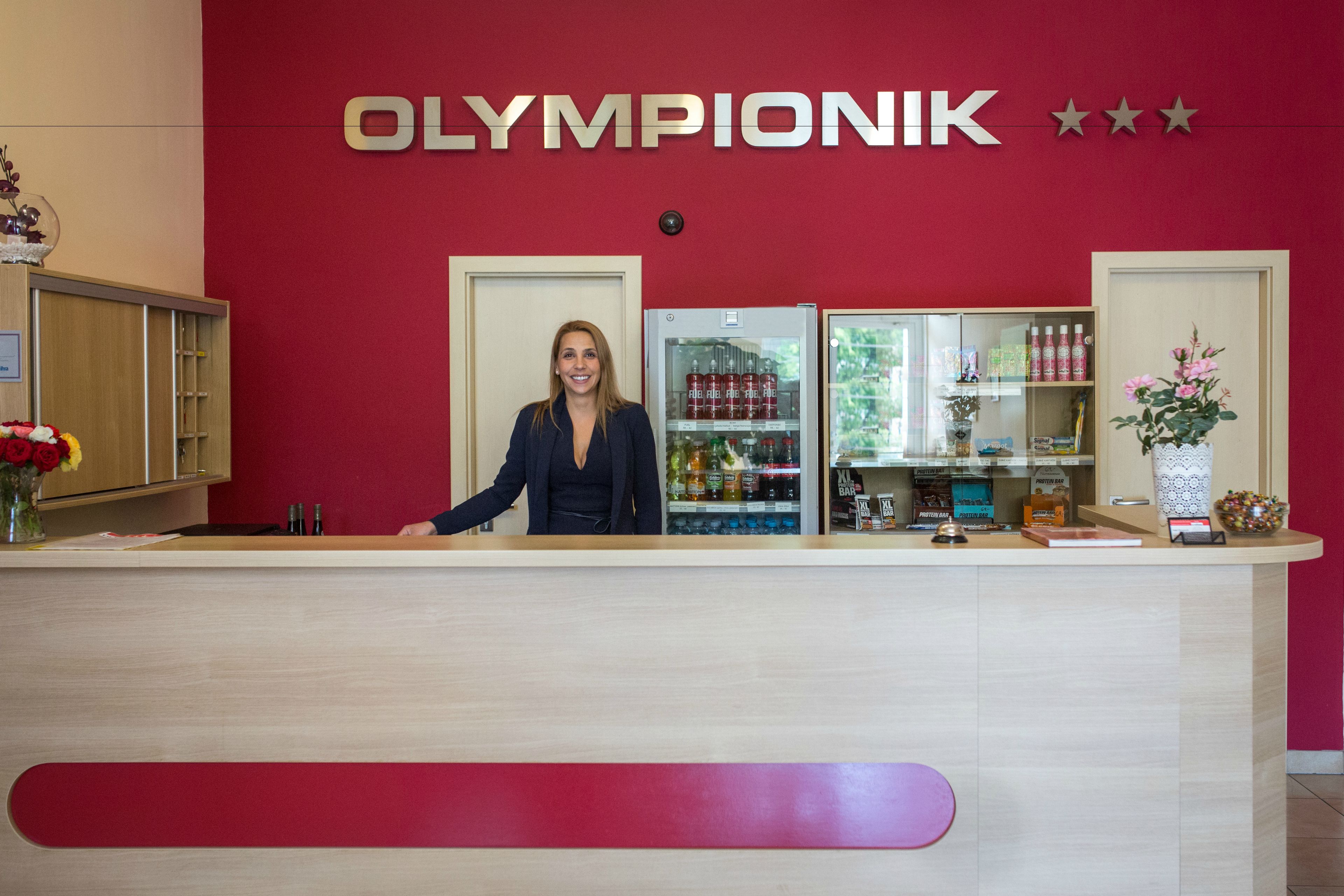 Un mostrador de recepción con un cartel que dice "OLYMPIONIK" y un refrigerador lleno de bebidas.
