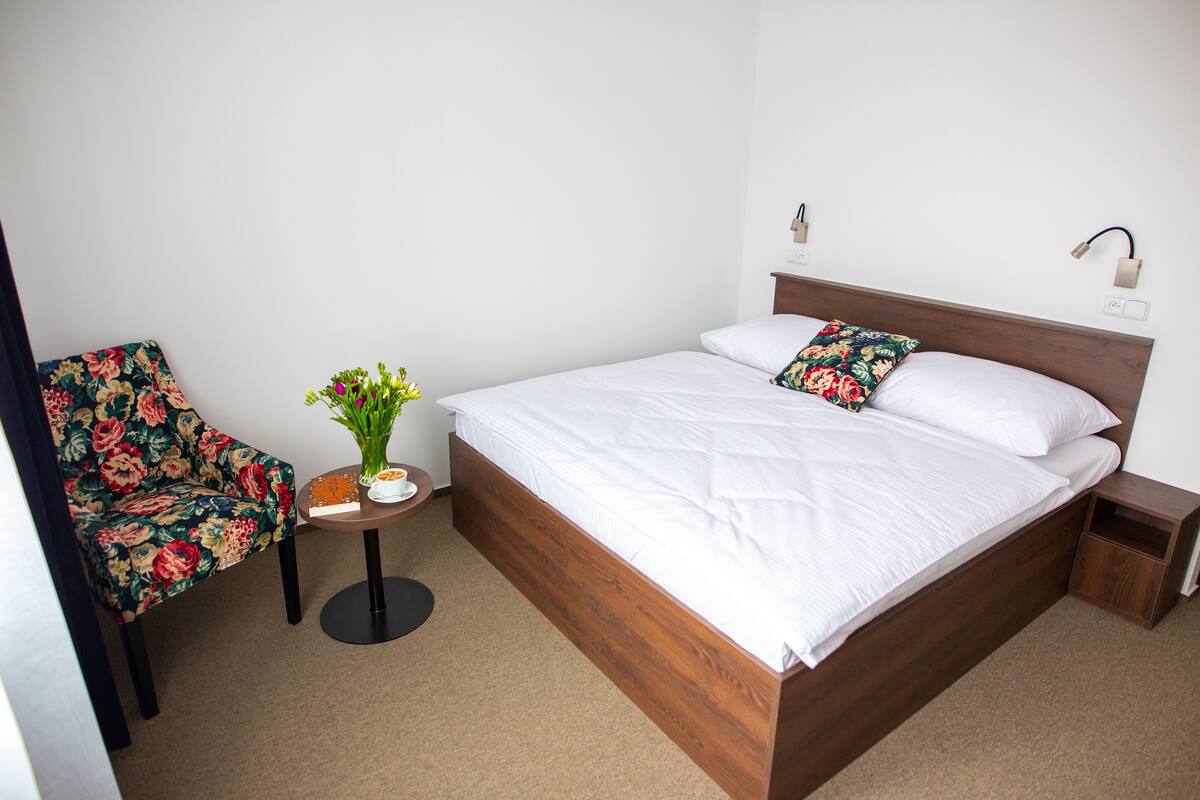 A neatly arranged bedroom with a wooden bed, a floral chair, and a small table with a vase of flowers.