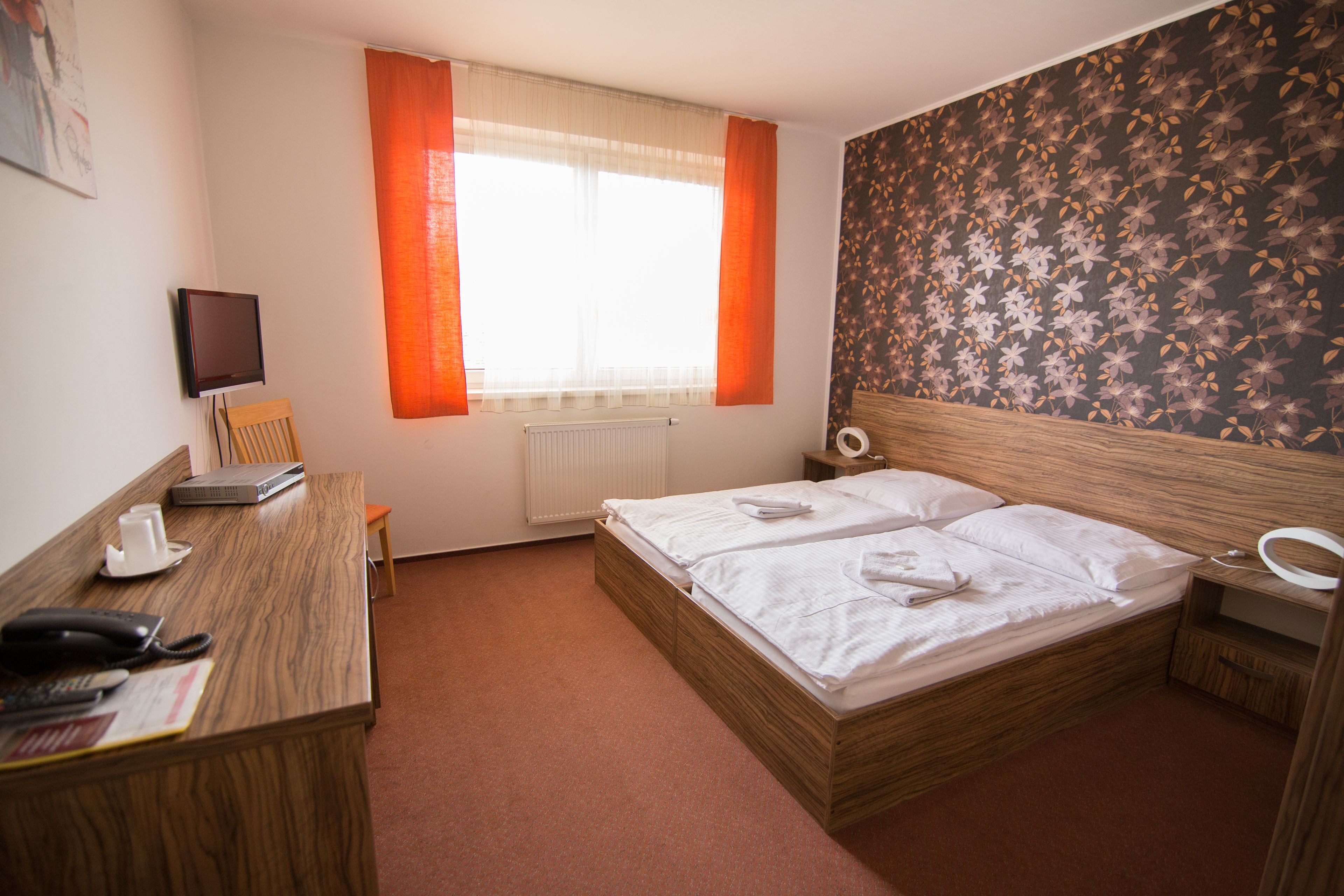 A hotel room with a wooden bed, a desk with a telephone and a cup, a radiator, and a television on the wall.
