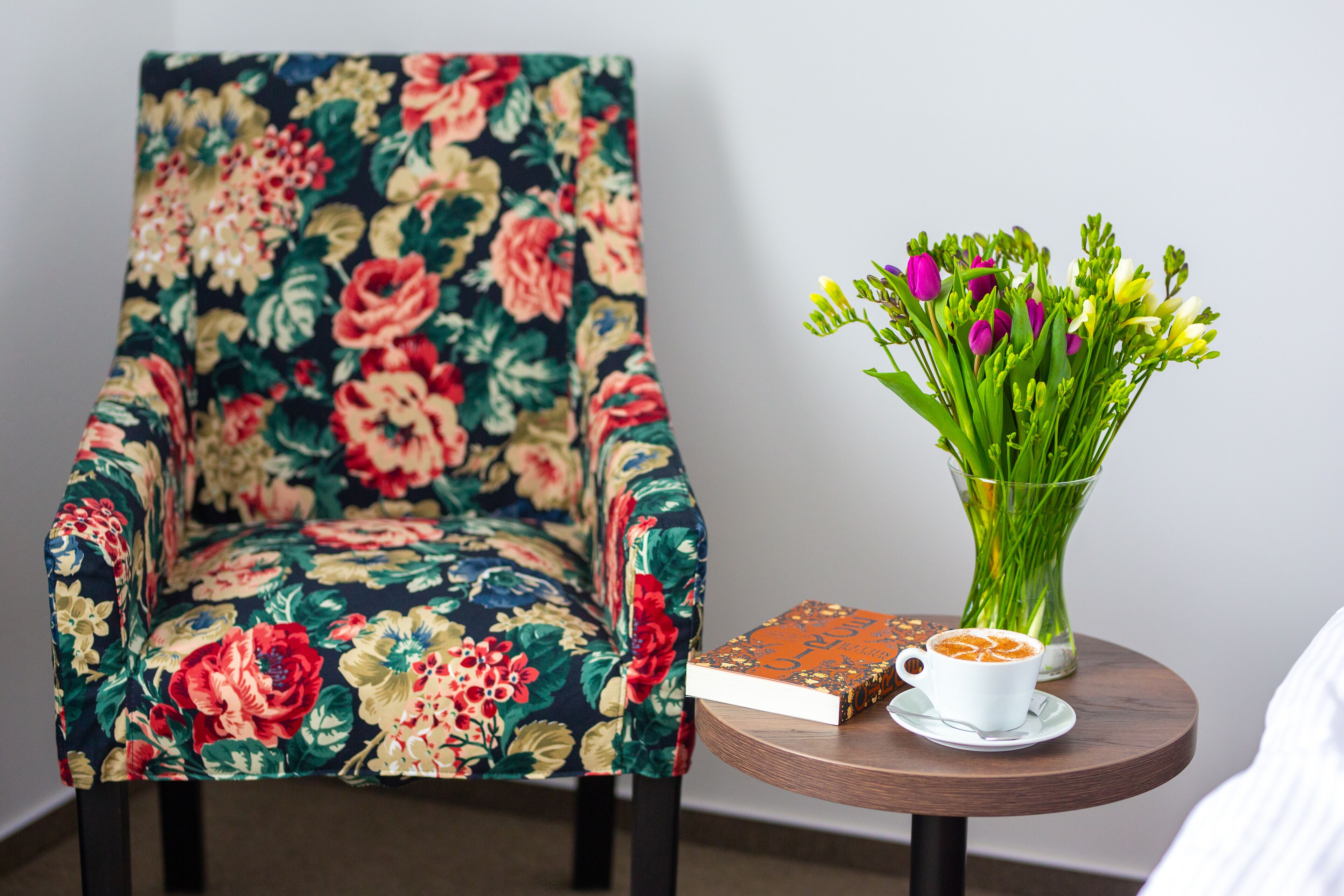 Ein floraler Sessel, ein Buch, eine Blumenvase und eine Kaffeetasse auf einem runden Holztisch.
