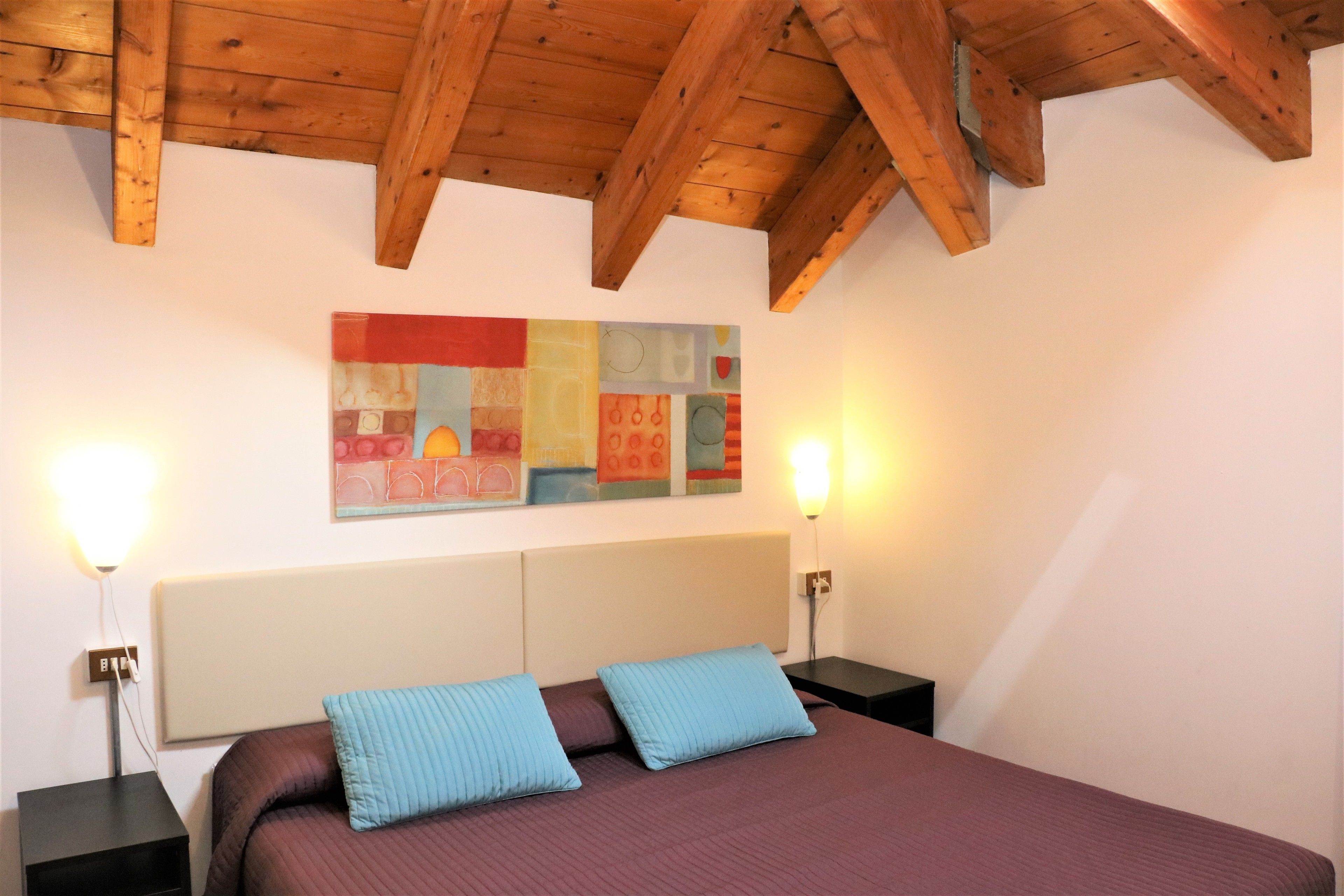 Ein Schlafzimmer mit Holzbalkendecke, einem großen Bett mit zwei Kissen, einem farbenfrohen abstrakten Gemälde an der Wand und zwei Nachttischlampen.