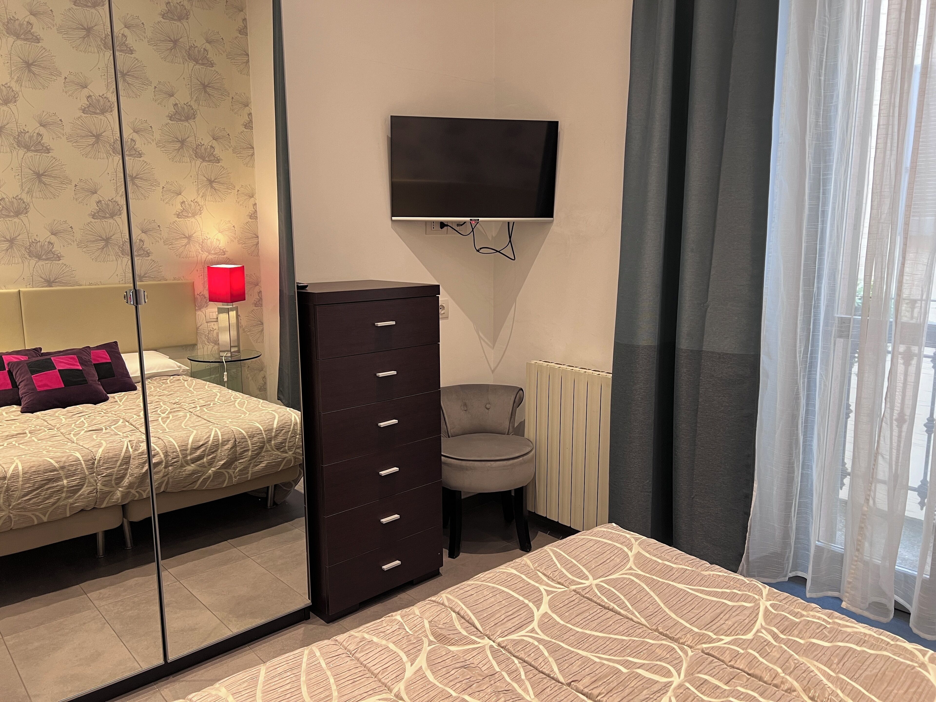 A hotel room with a bed, a dresser, a chair, a TV mounted on the wall, and a window with curtains.