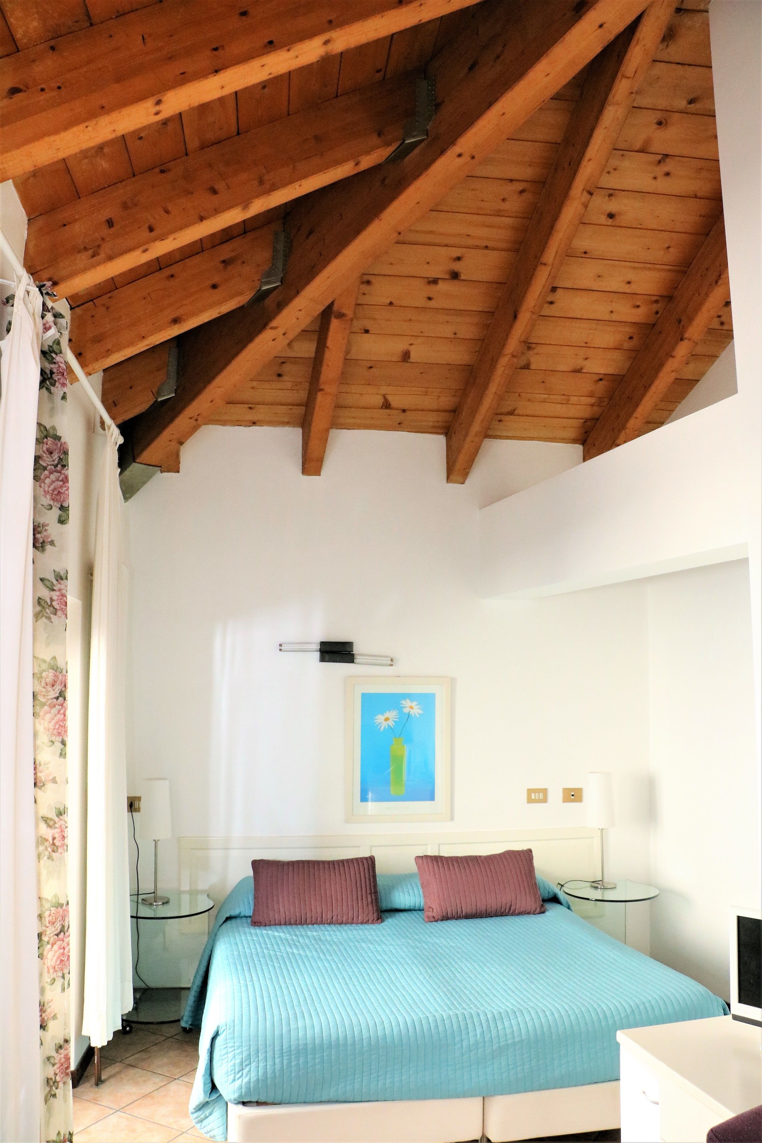 A bedroom with a wooden ceiling, a bed with blue bedding, a painting on the wall, and a bedside table with a lamp.