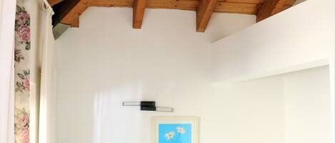 A bedroom with a wooden ceiling, a bed with blue bedding, a painting on the wall, and a bedside table with a lamp.