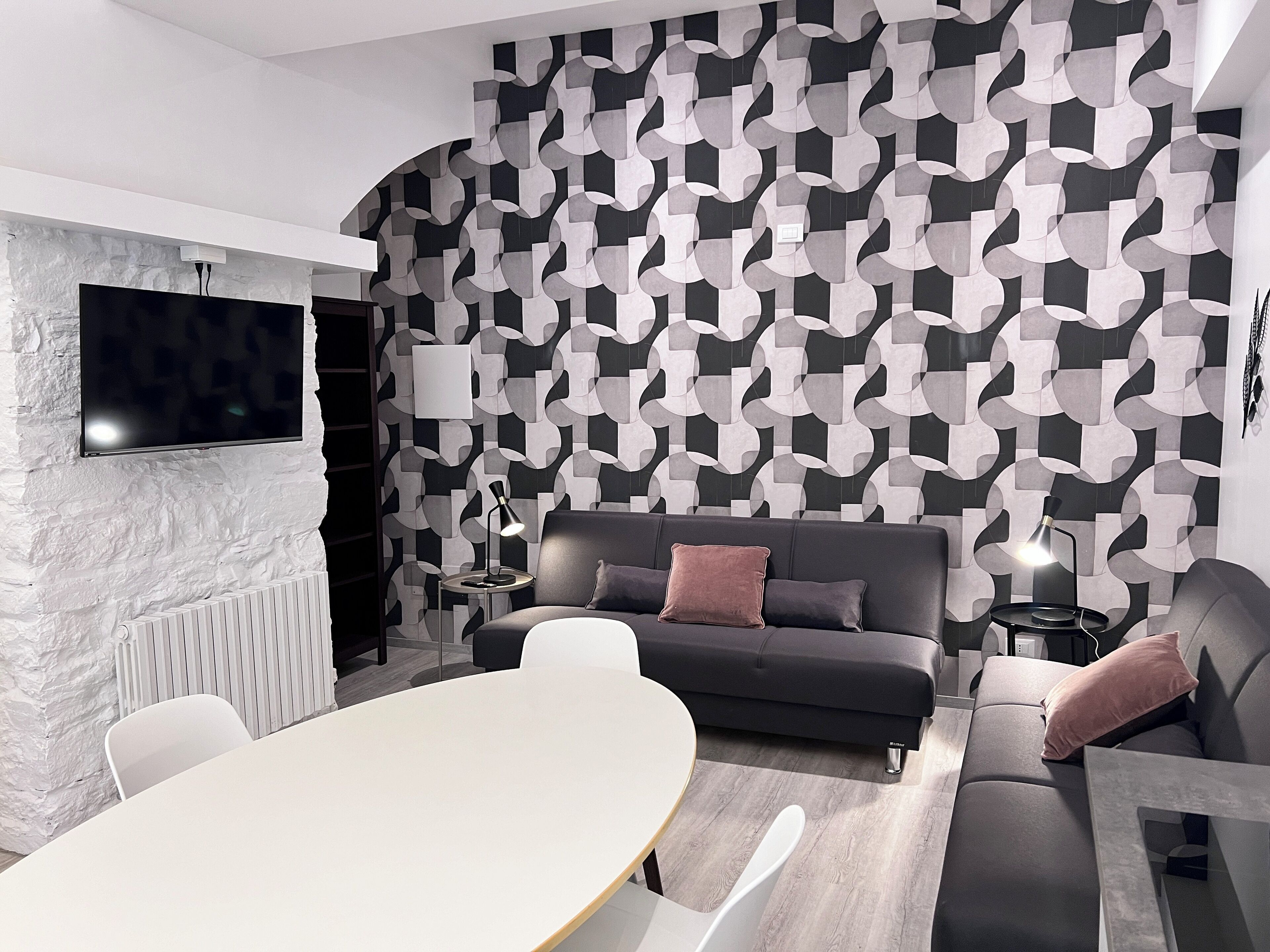 A modern living room with a geometric patterned wall, a flat-screen TV mounted on a textured wall, a dark grey sofa with cushions, a white round table, and a small side table with a lamp.