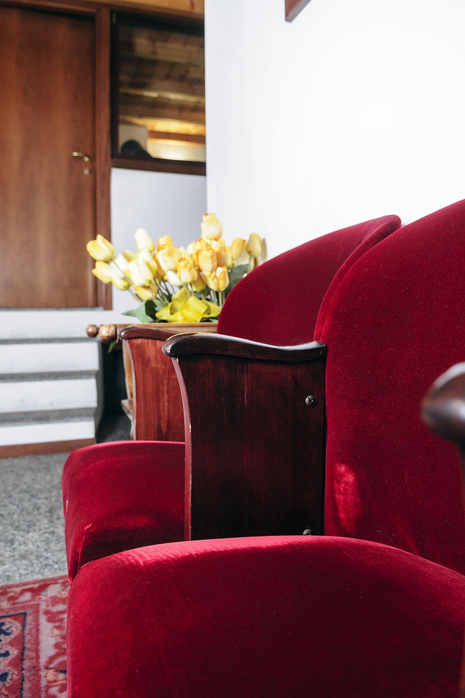Un sillón tapizado en terciopelo rojo con un reposabrazos de madera y un florero con flores amarillas sobre una mesa de madera.