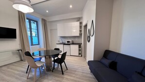 A modern living room with a dark blue sofa, a glass dining table with four chairs, and a kitchen area with white cabinets and a microwave. - Residence Diaz (Como)