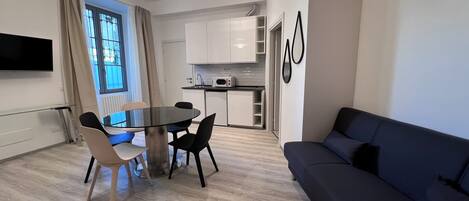 A modern living room with a dark blue sofa, a glass dining table with four chairs, and a kitchen area with white cabinets and a microwave.