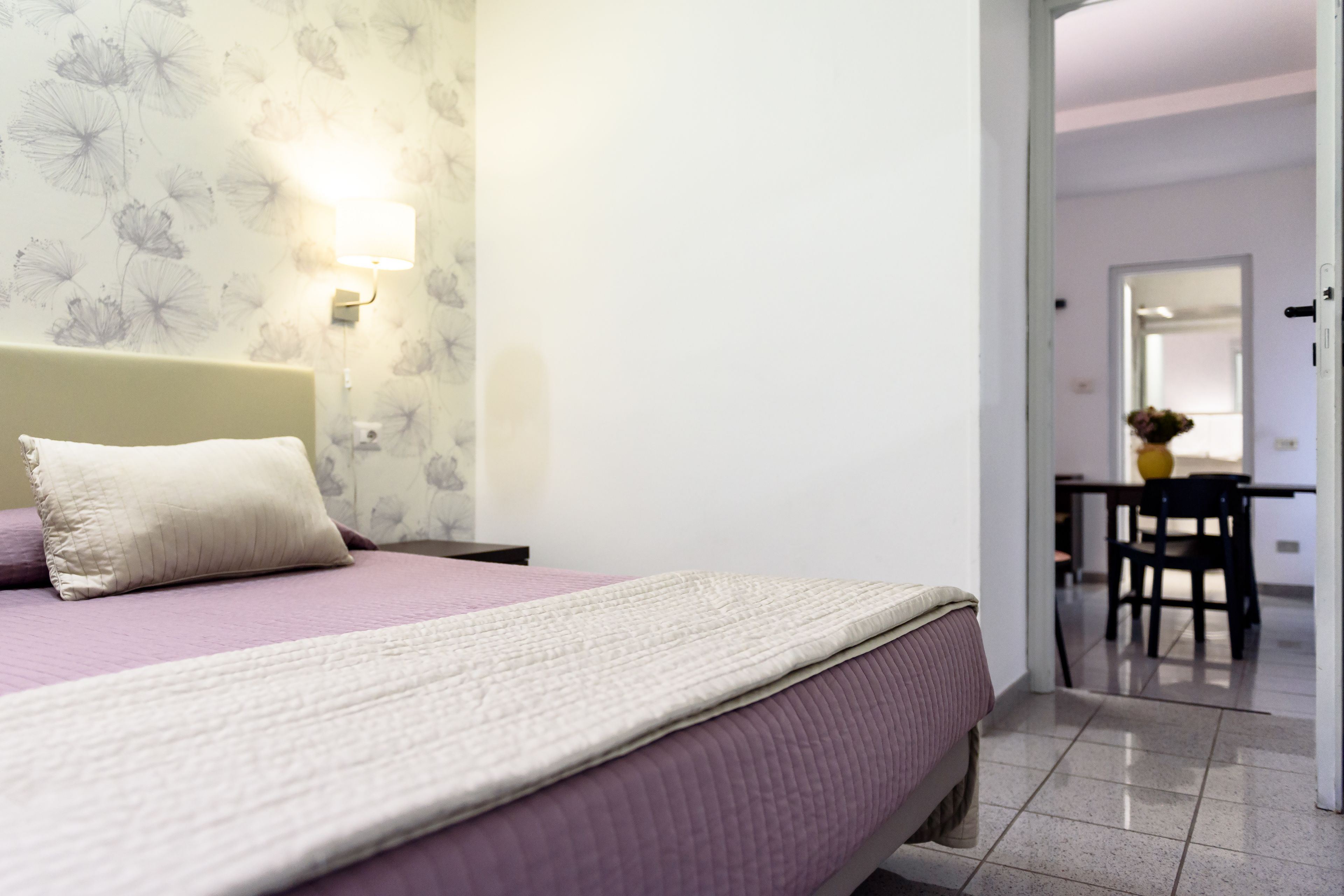 Ein Schlafzimmer mit Bett, Nachttisch, Lampe und Blick zum Essbereich.
