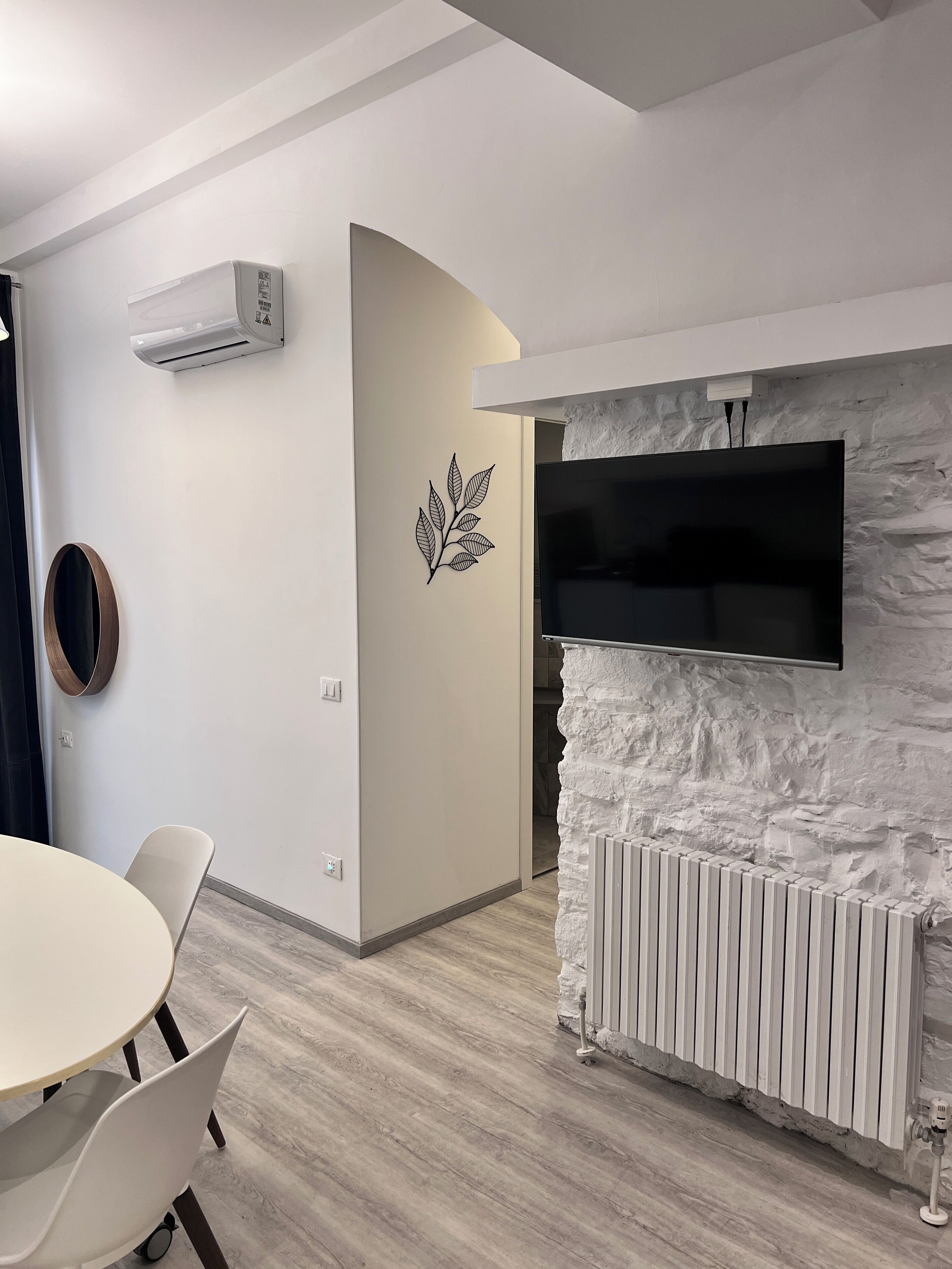 Ein modernes Zimmer mit weißer Wand, einer Wand-Klimaanlage, einem Flachbildfernseher, einem runden Spiegel und einem Esstisch mit Stühlen.