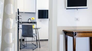 Habitación de hotel con una cama, un televisor en la pared, una pequeña mesa de madera y una kitchenette al fondo.