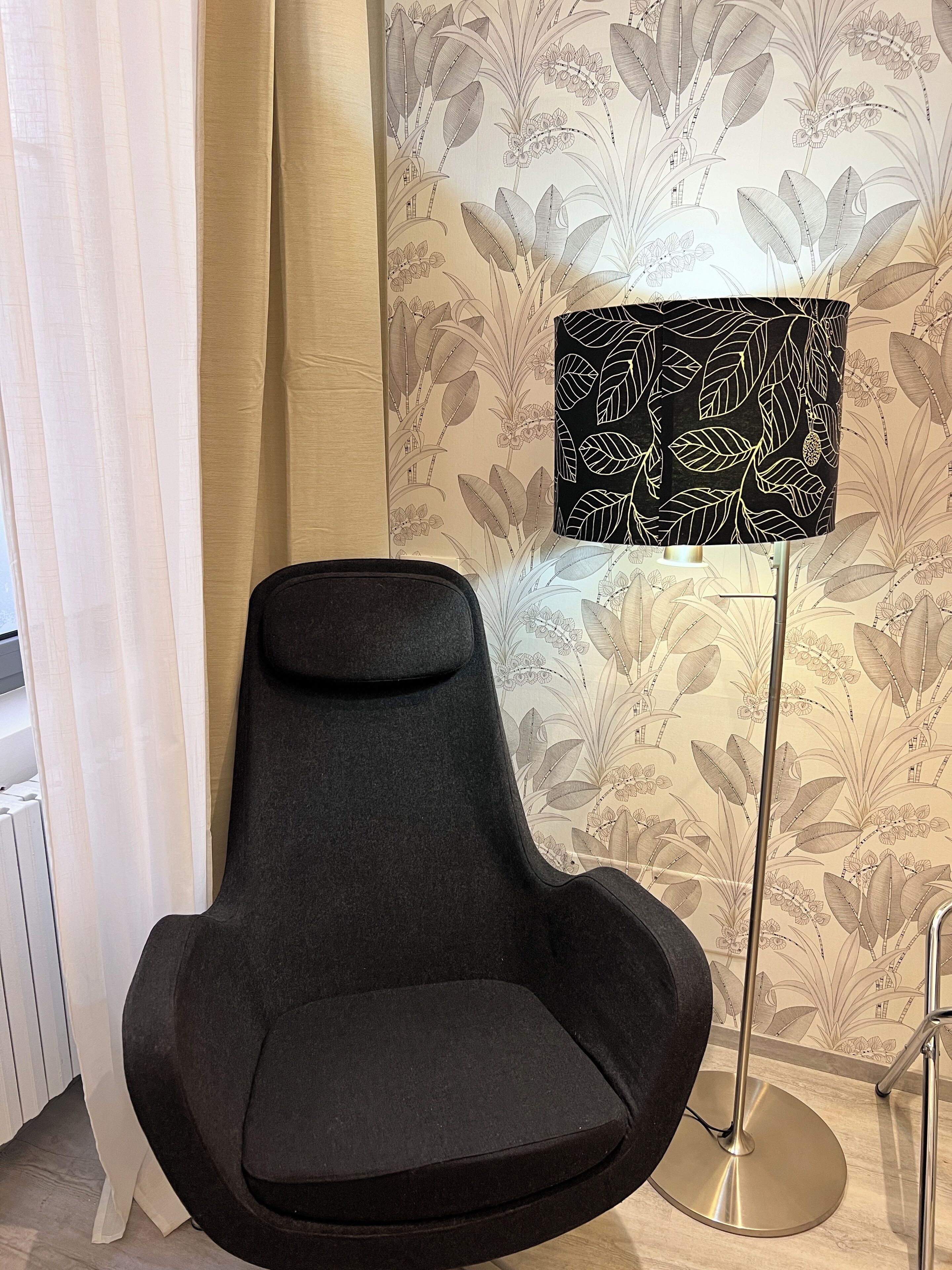 A modern room with a black armchair, a floor lamp with a black and white leaf pattern, and floral wallpaper.