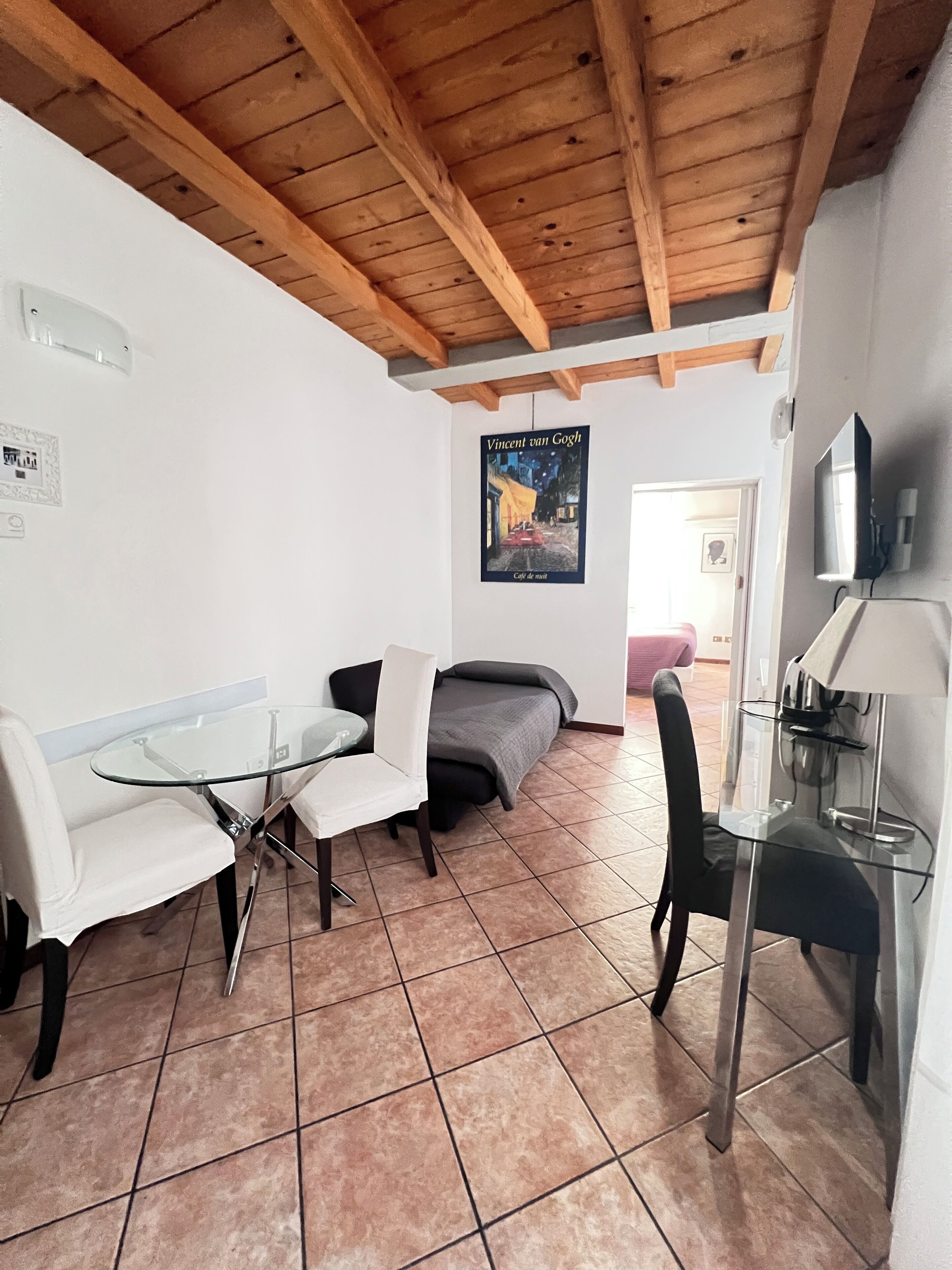 A room with a wooden ceiling, a glass table with chairs, a sofa, a poster on the wall, and a TV mounted on the wall.
