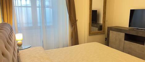 A hotel room with a bed, a TV on a stand, a mirror, and a window with sheer curtains.