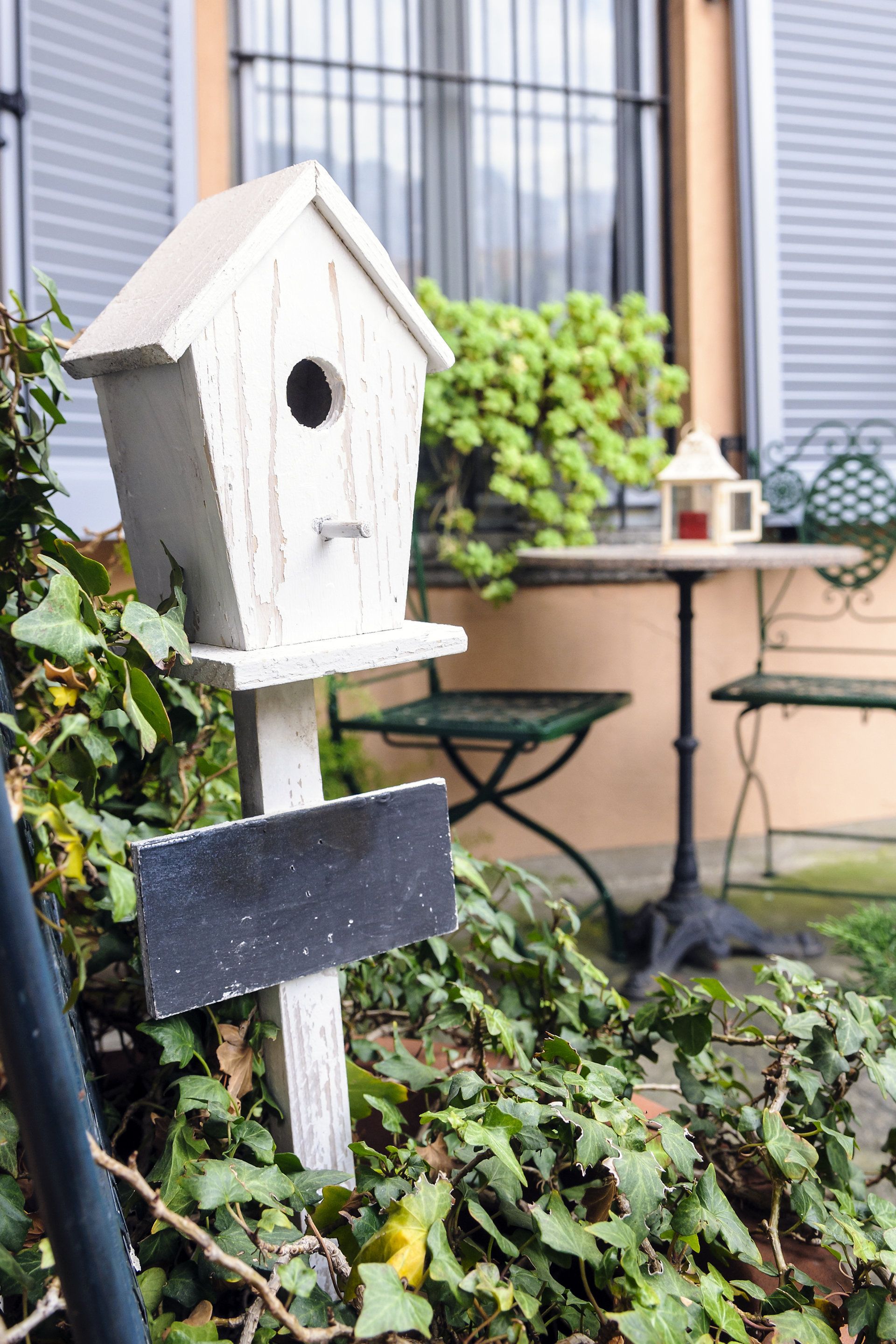 Un comedero para pájaros blanco sujeto a un poste con un letrero negro, rodeado de follaje.