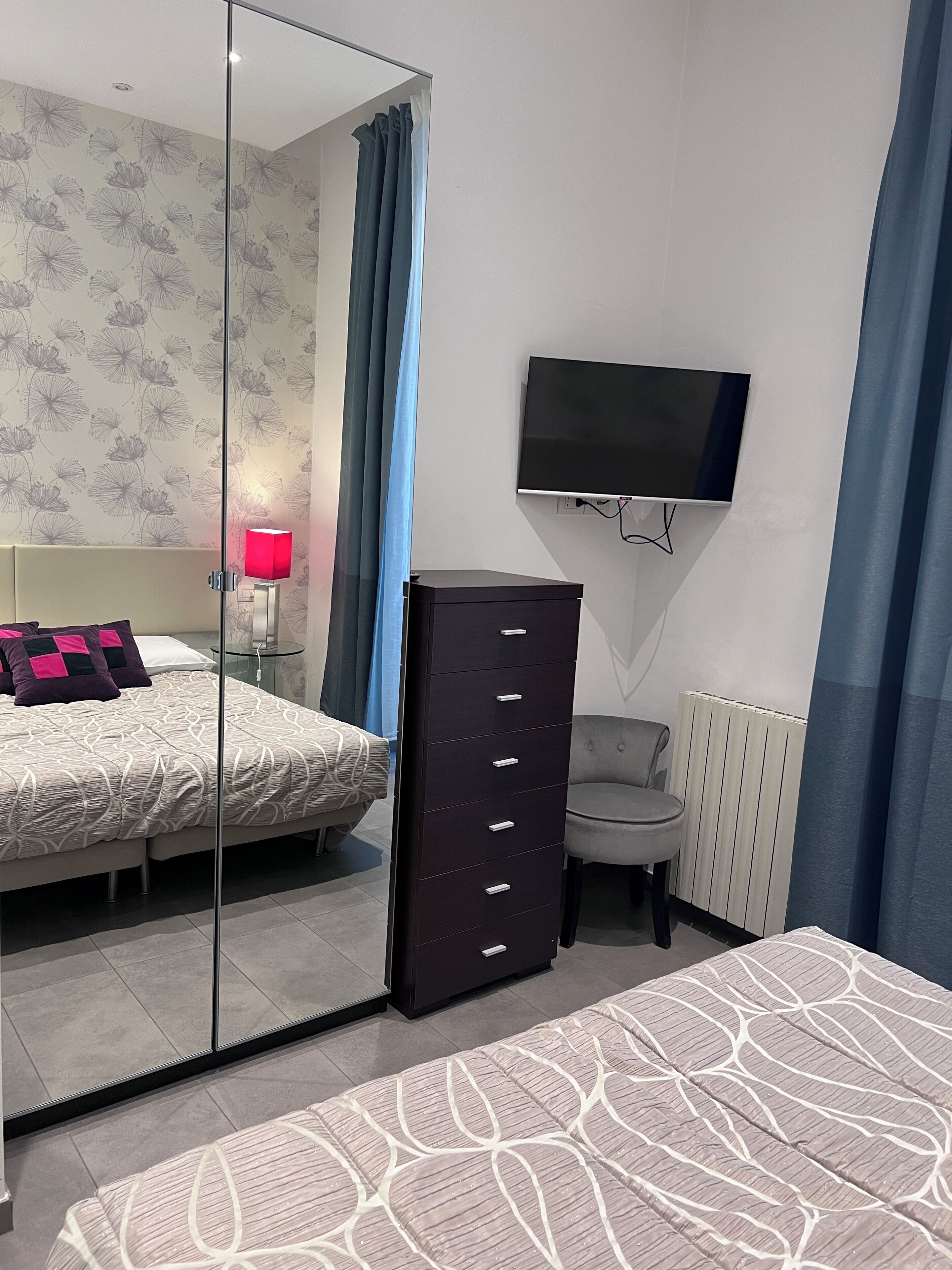 Quarto moderno com um espelho grande, cama com roupa de cama estampada, cômoda, cadeira e televisão fixada na parede.