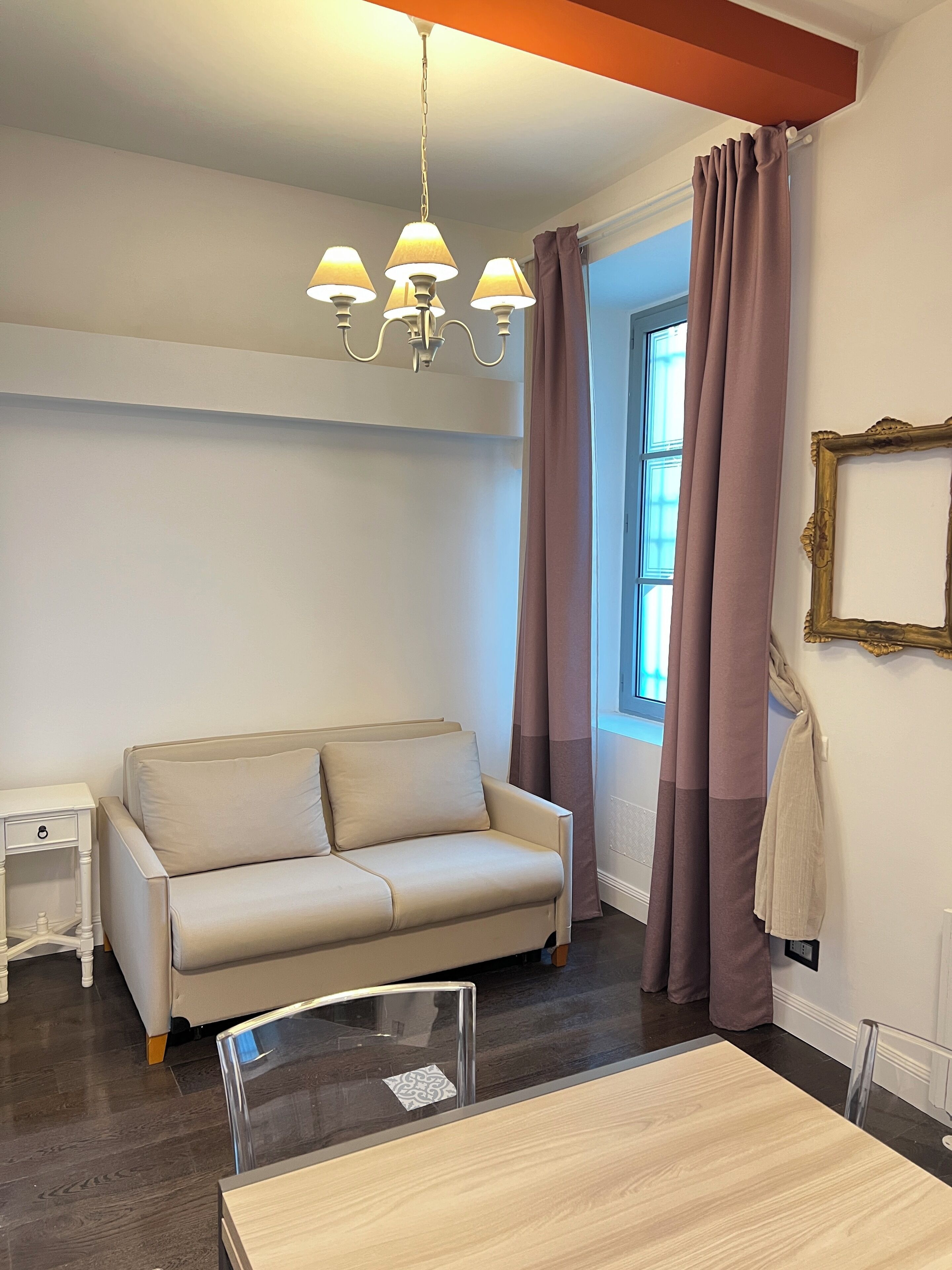 A room with a beige sofa, a small white nightstand, a table with a chair, a chandelier, and a window with curtains.