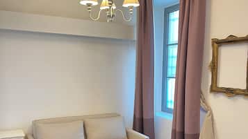 A room with a beige sofa, a small white nightstand, a table with a chair, a chandelier, and a window with curtains.