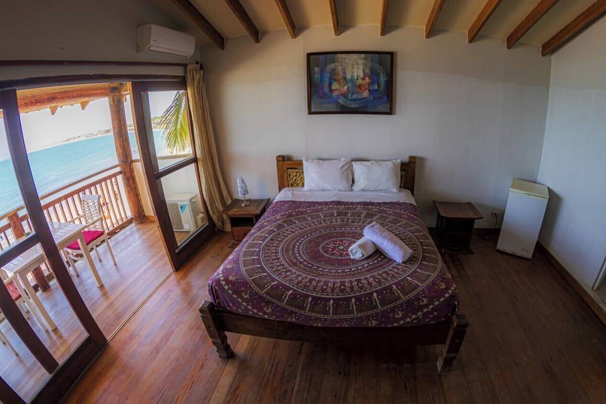 A bedroom with a wooden bed, a painting on the wall, and a view of the sea through a balcony.