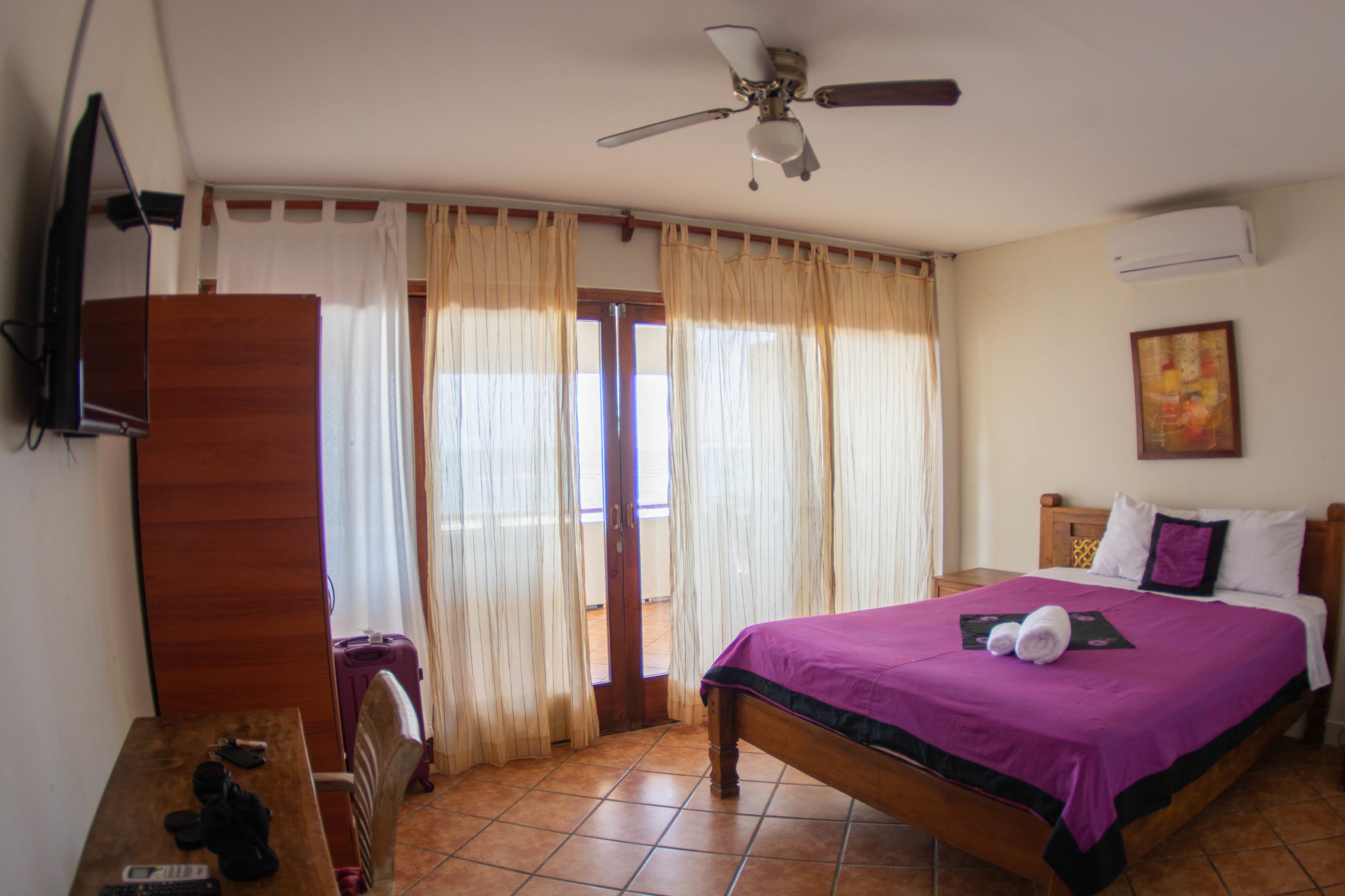 Un dormitorio con cama, cortinas, ventilador de techo, cómoda de madera y televisor en la pared.