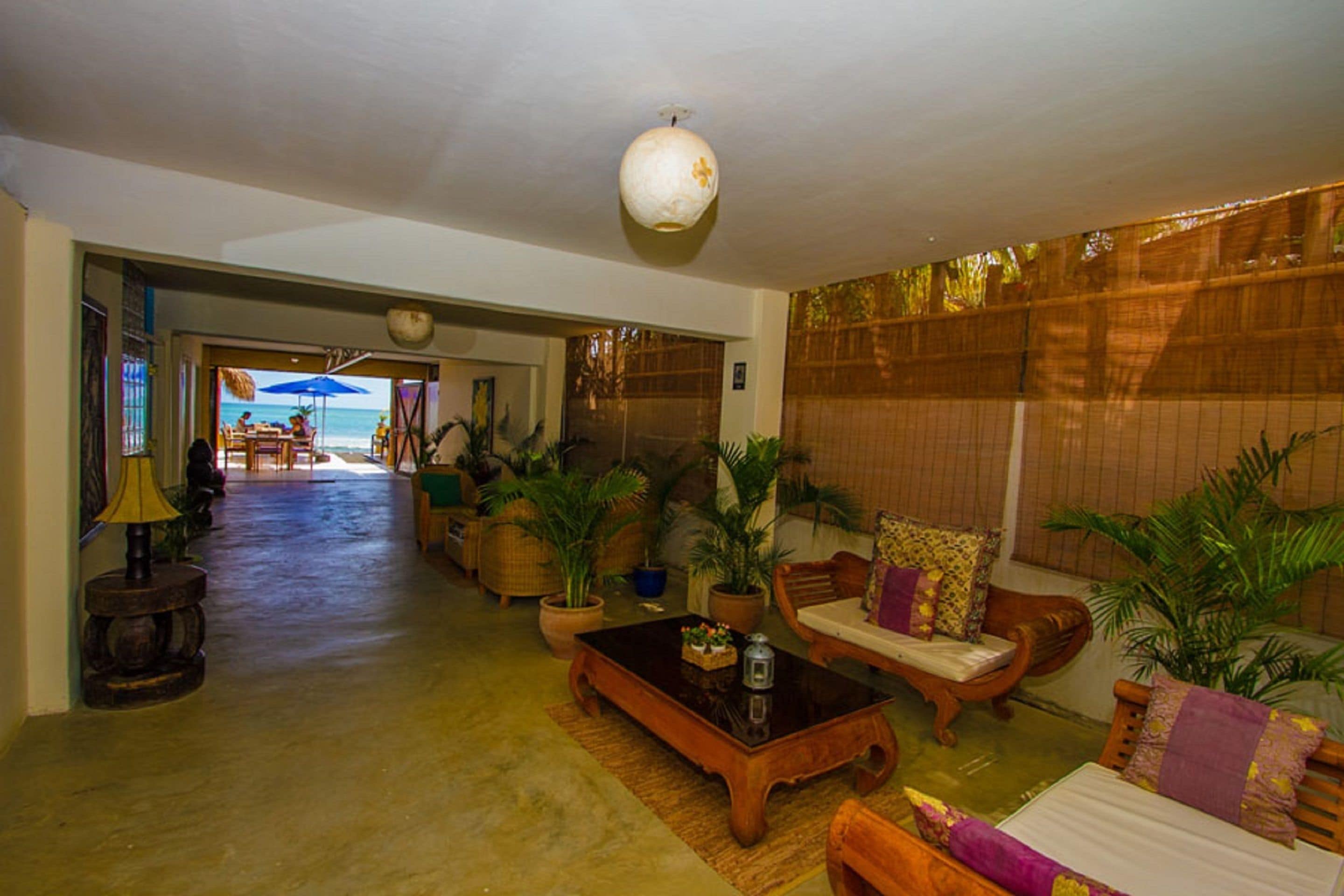 A spacious living area with a large wooden coffee table, several sofas, and potted plants. There is a view of the ocean through a doorway.