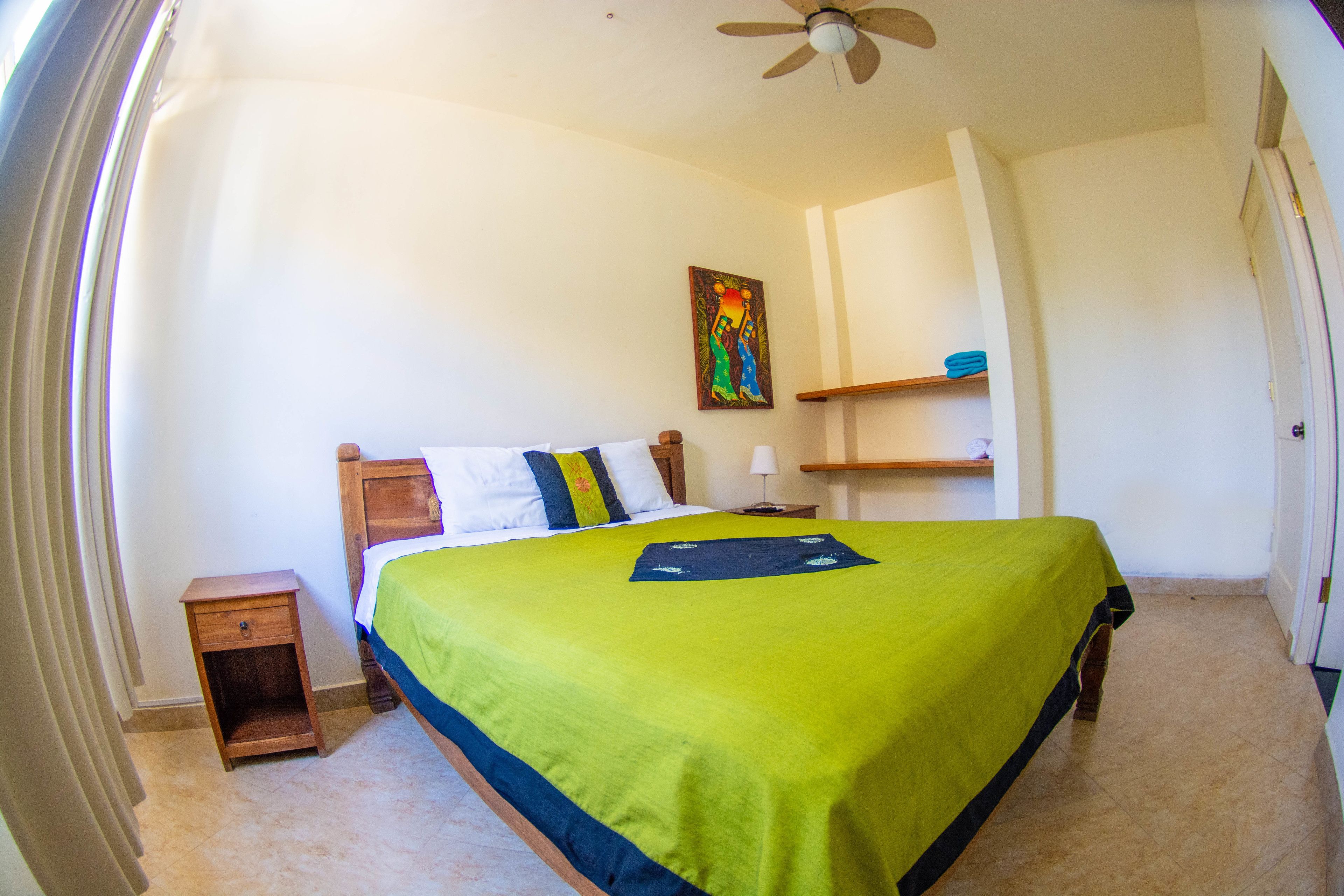 A bedroom with a bed, a nightstand, a ceiling fan, a painting on the wall, and a shelf with a bowl.