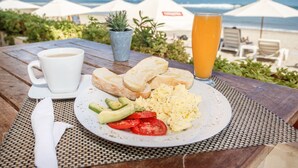 A breakfast setting with a plate of scrambled eggs, toast, avocado, and tomatoes, a cup of coffee, and a glass of orange juice, overlooking a beach with umbrellas. - Don Giovanni Máncora (Máncora)