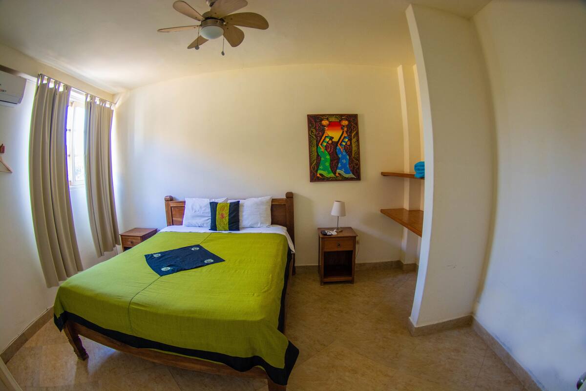 A hotel room with a bed, a ceiling fan, a painting on the wall, and a nightstand with a lamp.