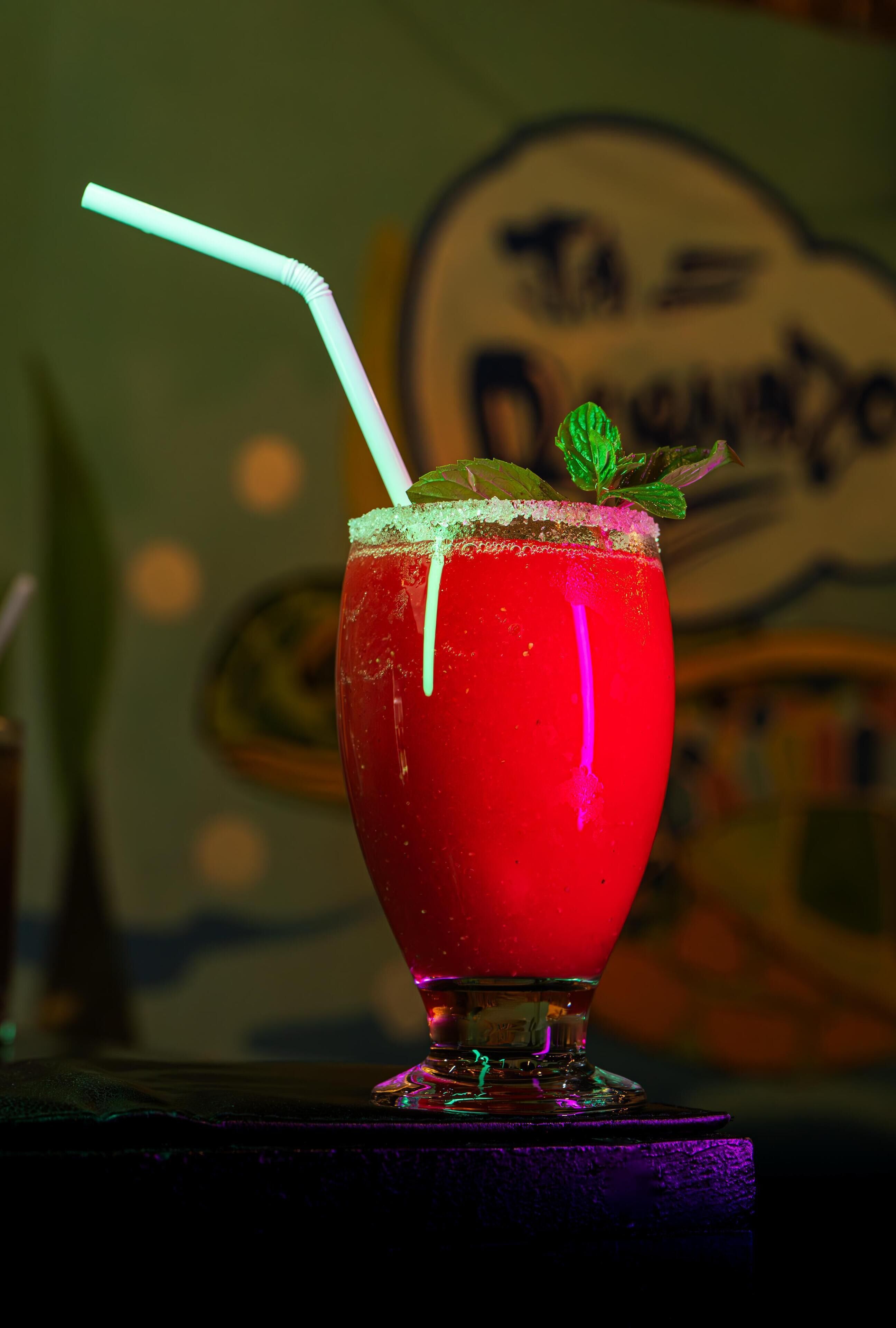 A glass of red beverage with a straw and mint garnish, placed on a table with a blurred background featuring a sign.