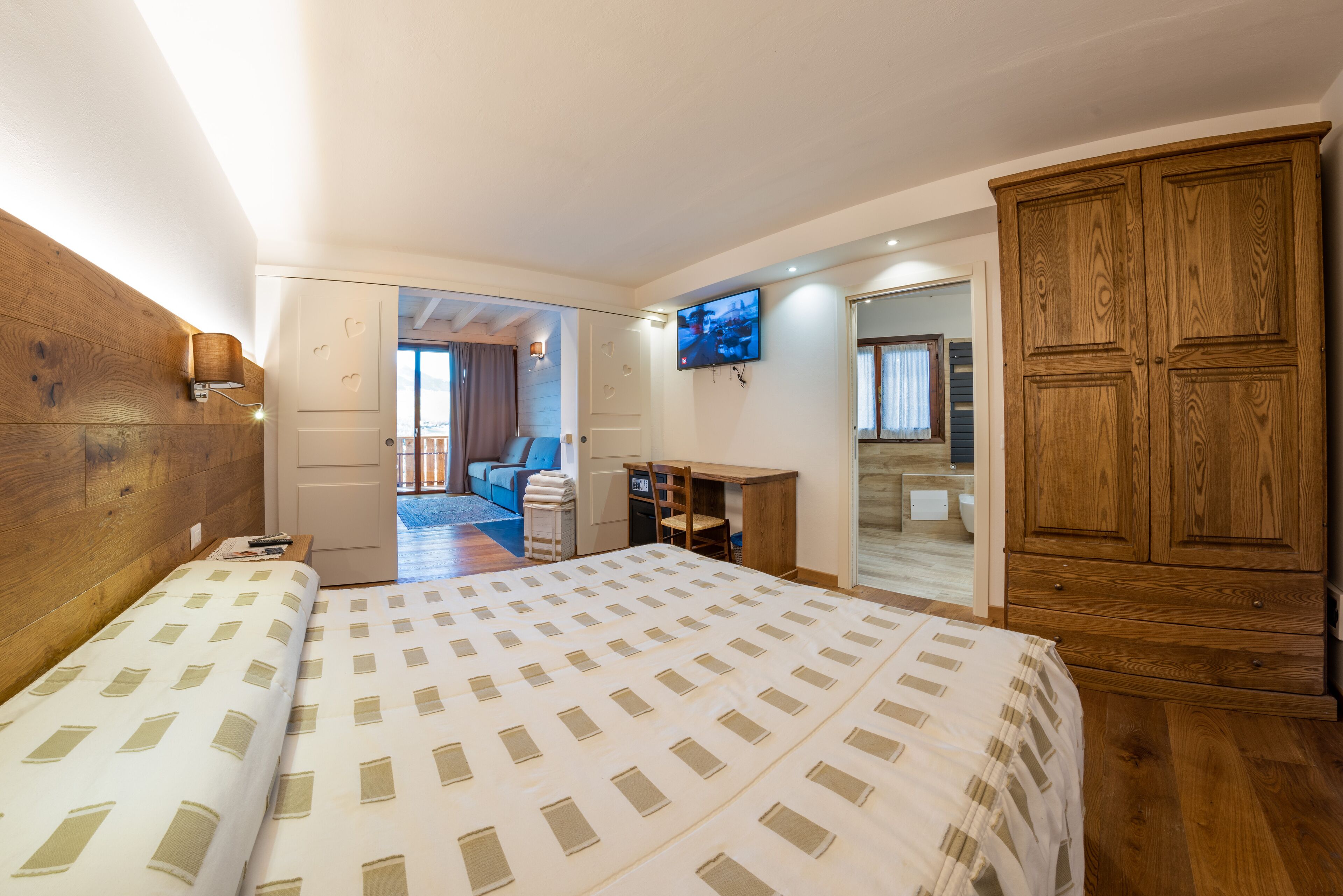 A bedroom with a large bed, wooden wardrobe, and a TV on the wall.