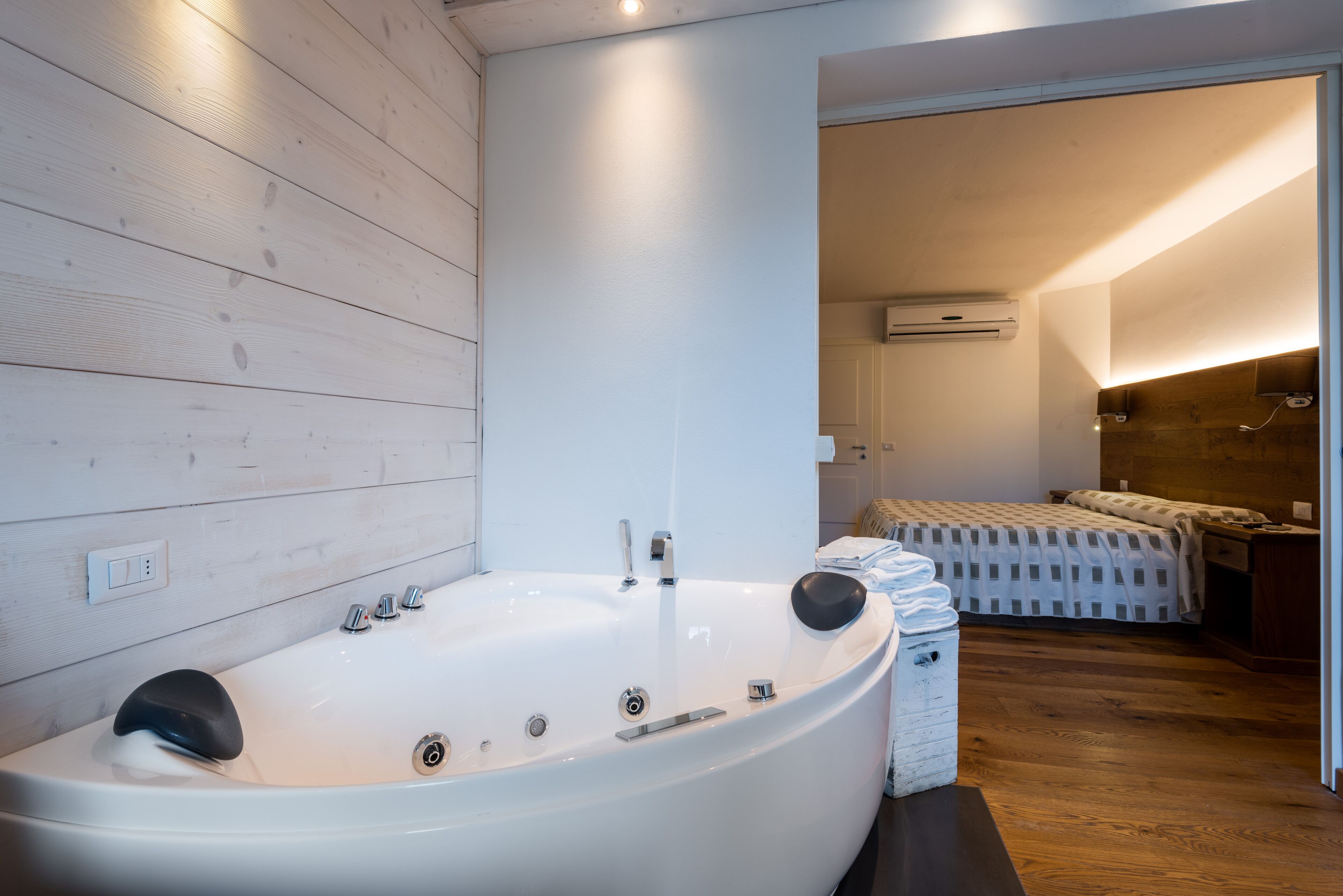 Un baño moderno con una bañera grande, paneles de madera en las paredes, un equipo de aire acondicionado a la vista y un dormitorio con cama y mesita de noche.
