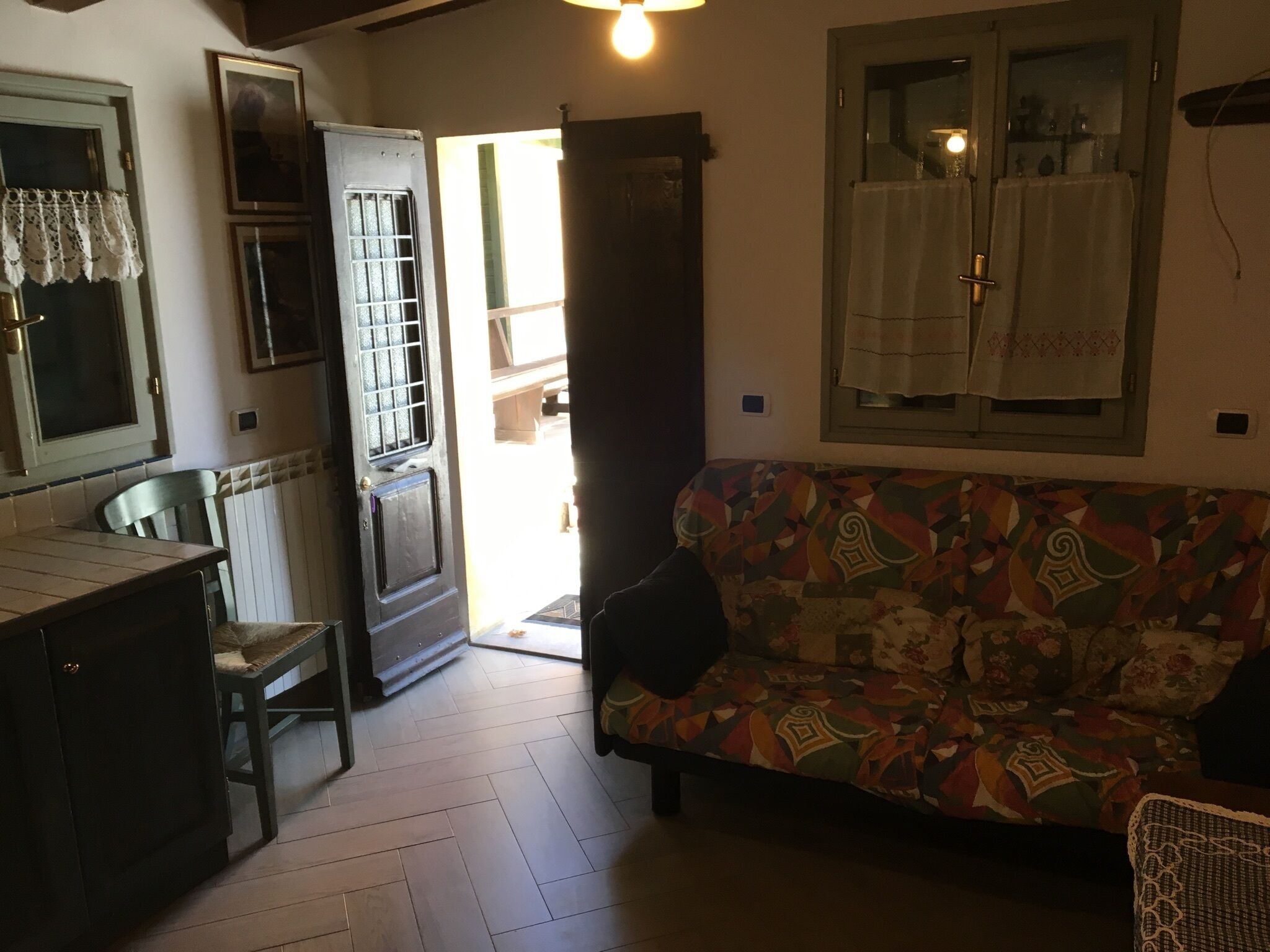 A cozy living room with a patterned sofa, a dining table with chairs, and a kitchen area with a refrigerator and a window with lace curtains.