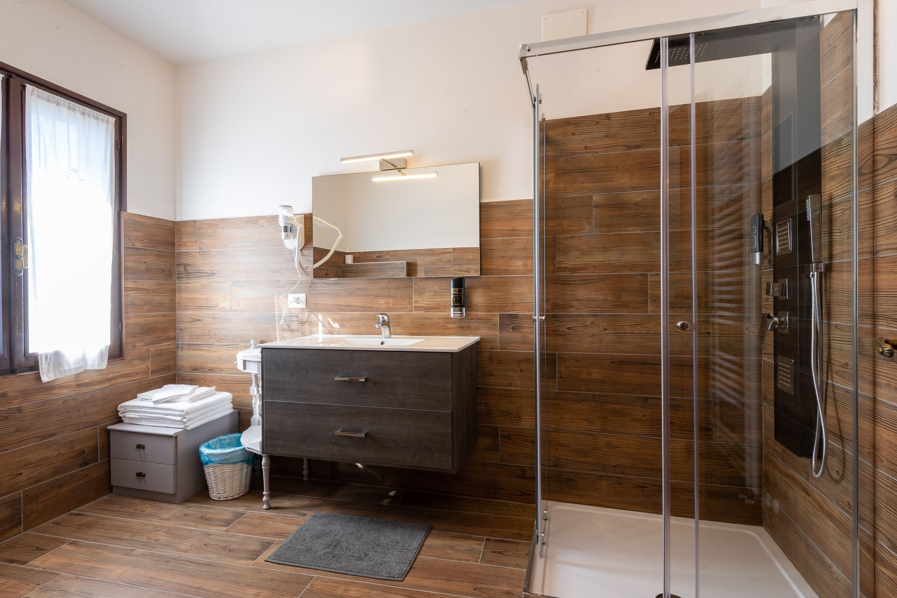 A modern bathroom with a wooden floor, a glass shower enclosure, a sink with a mirror, and a small bedside table with a trash bin.