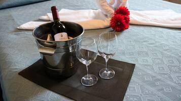 Un letto con una coperta azzurra a motivi decorativi, un asciugamano bianco, un fiore rosso, una bottiglia di vino e due calici vuoti su un sottopiattino scuro.