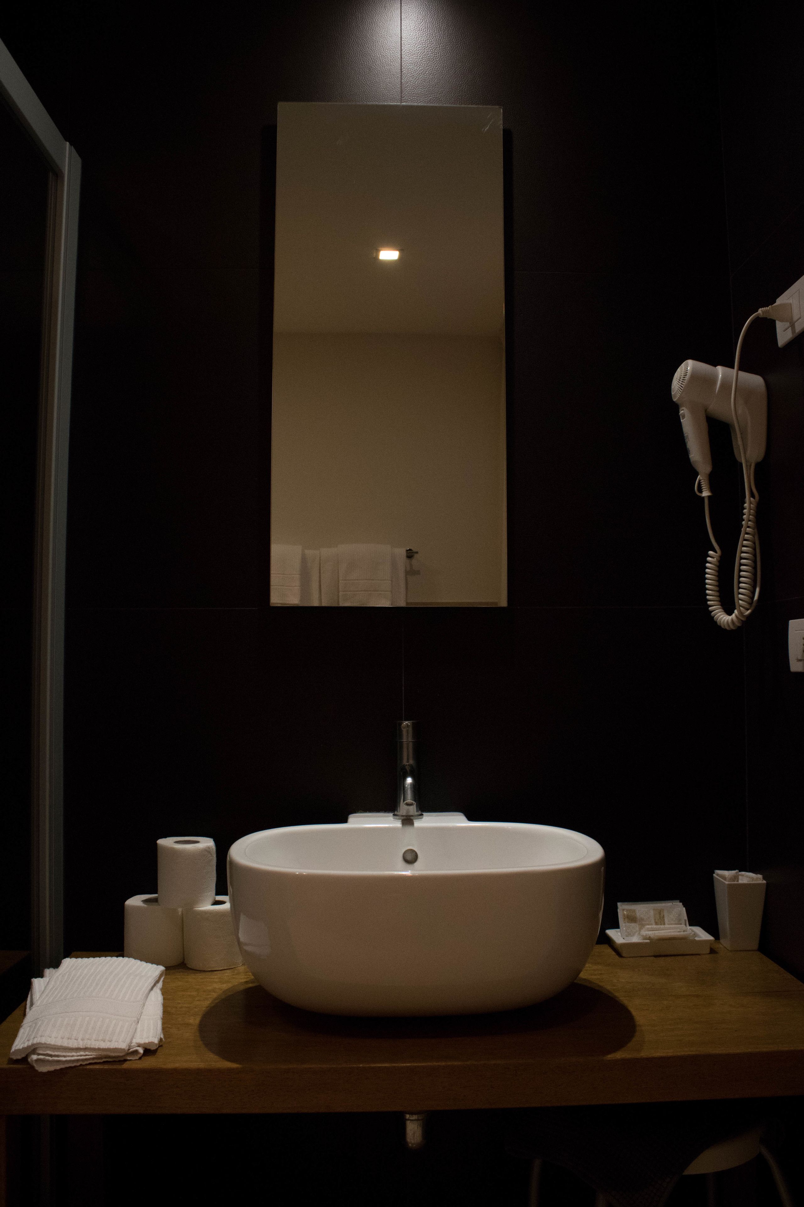 A bathroom with a large white sink, a mirror above it, a hairdryer on the wall, and a towel on the counter.