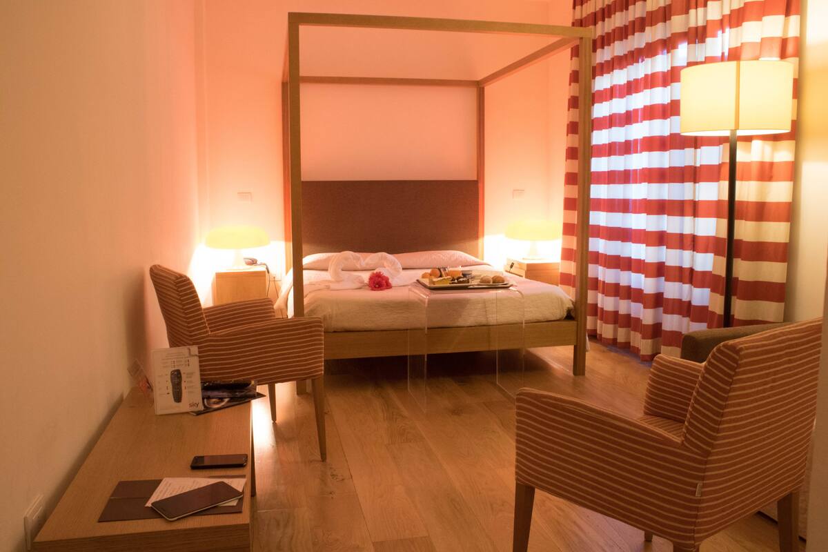 A four-poster bed with a wooden headboard, two bedside lamps, a striped curtain, and two striped armchairs.