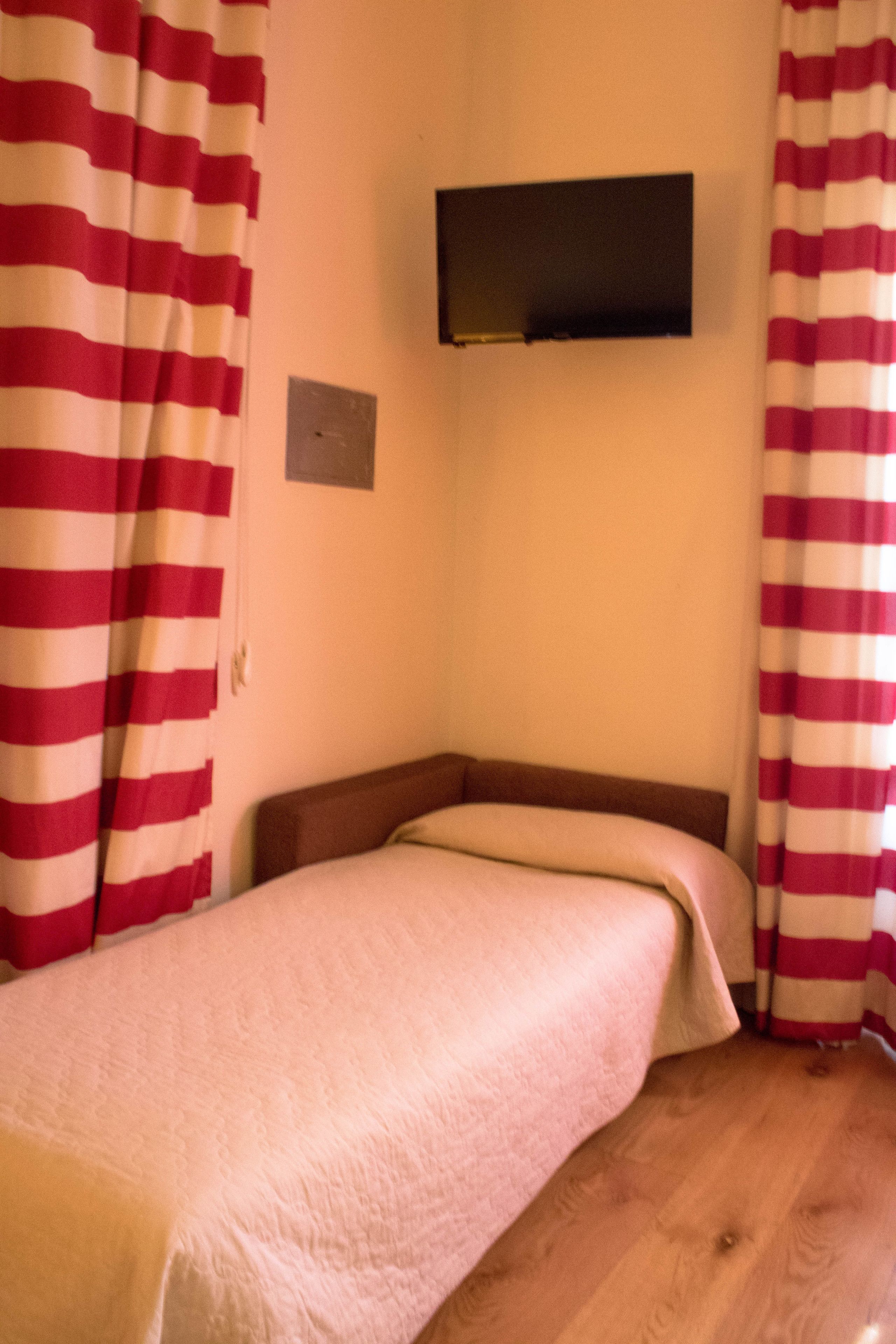 Una habitación pequeña con cama individual, suelo de madera, televisor de pantalla plana montado en la pared y cortinas a rayas.