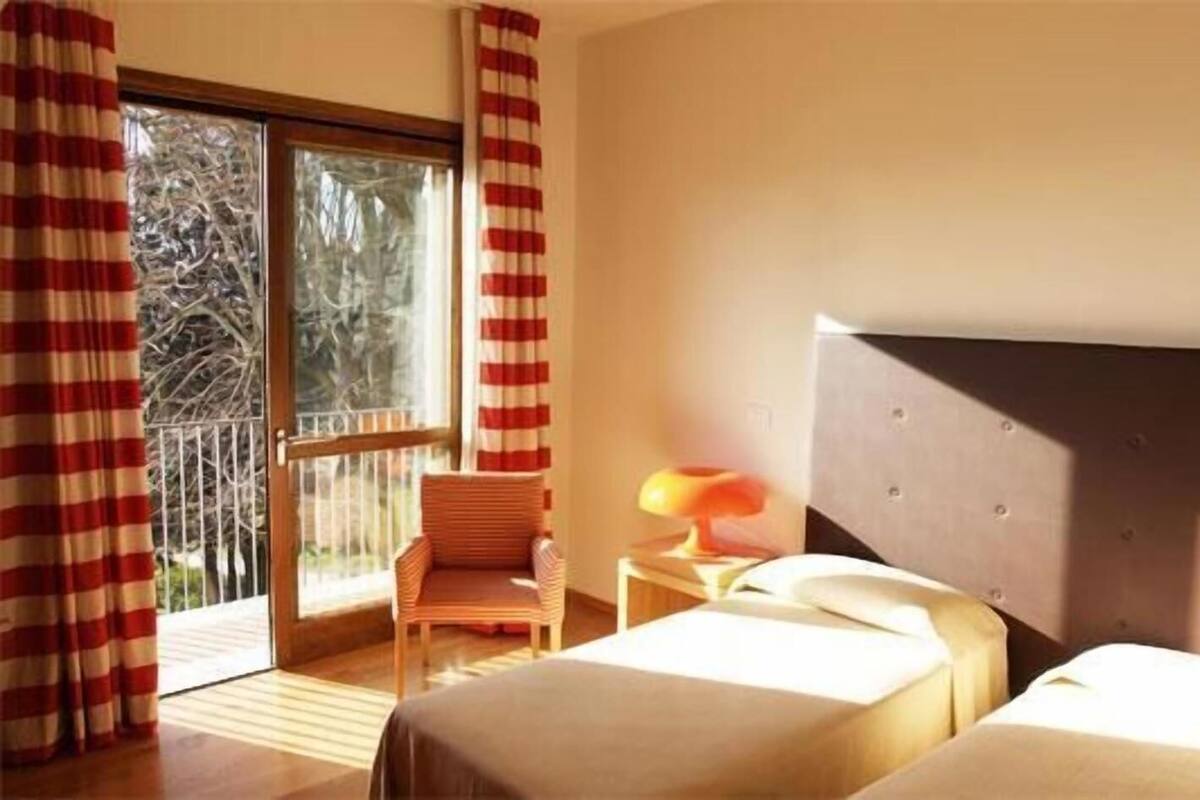 A hotel room with two beds, a chair, a table, and a balcony with curtains.
