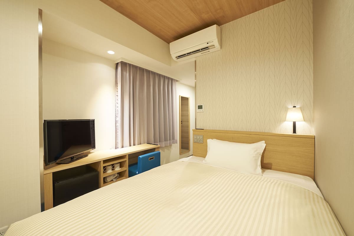 A hotel room with a bed, a desk with a television, a desk lamp, and an air conditioning unit.