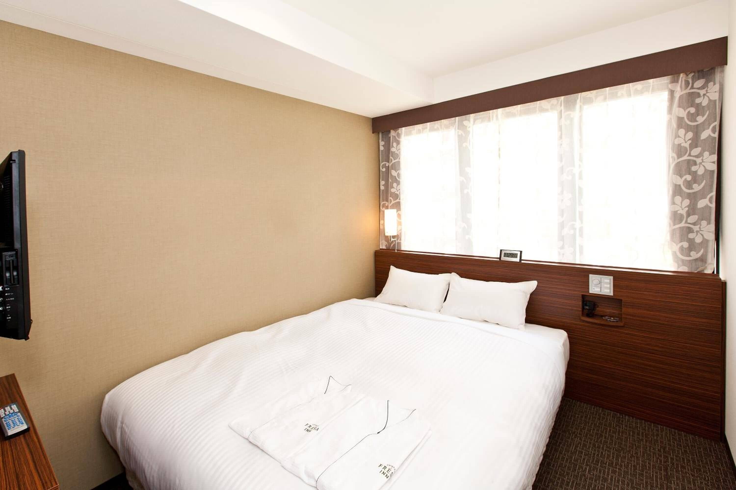 Ein Hotelzimmer mit einem großen Bett, einem hölzernen Kopfteil, einem an der Wand montierten Fernseher und einem Fenster mit floralen Vorhängen.
