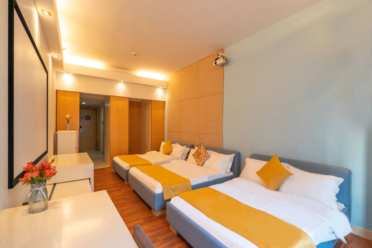 Habitación de hotel con tres camas, un escritorio y una mesita con un jarrón de flores.