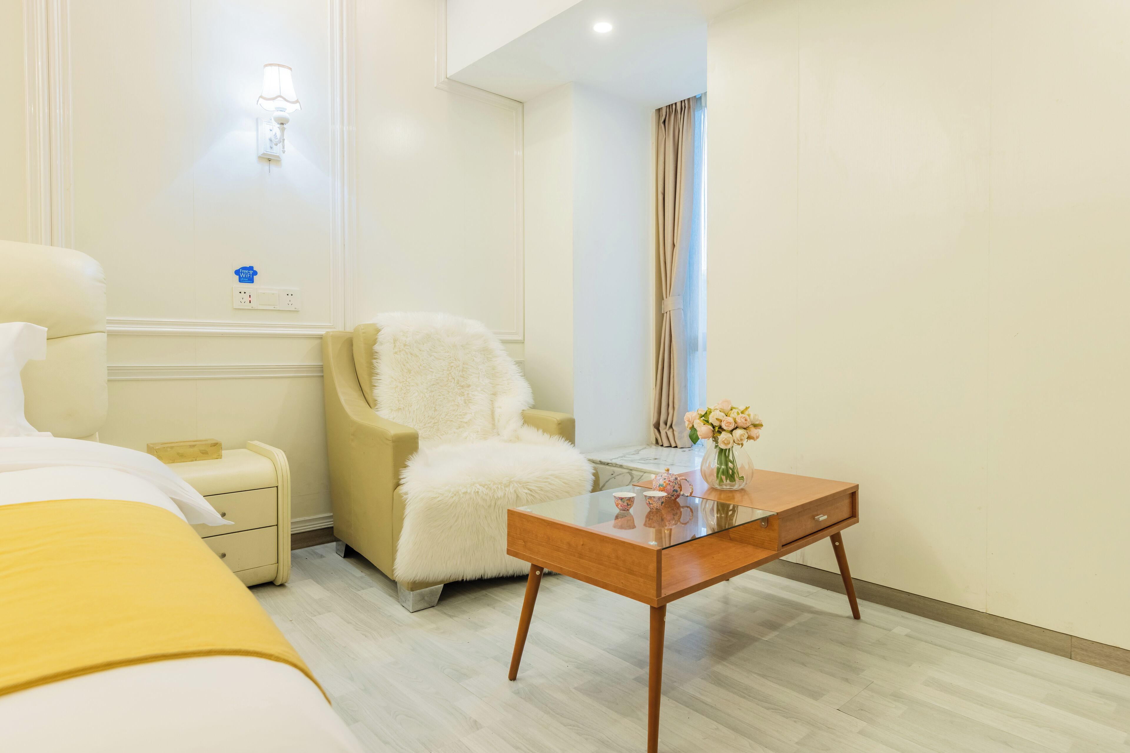 Een hotelkamer met een bed, een comfortabele fauteuil, een klein tafeltje met een bloemenvaas en een nachtkastje.