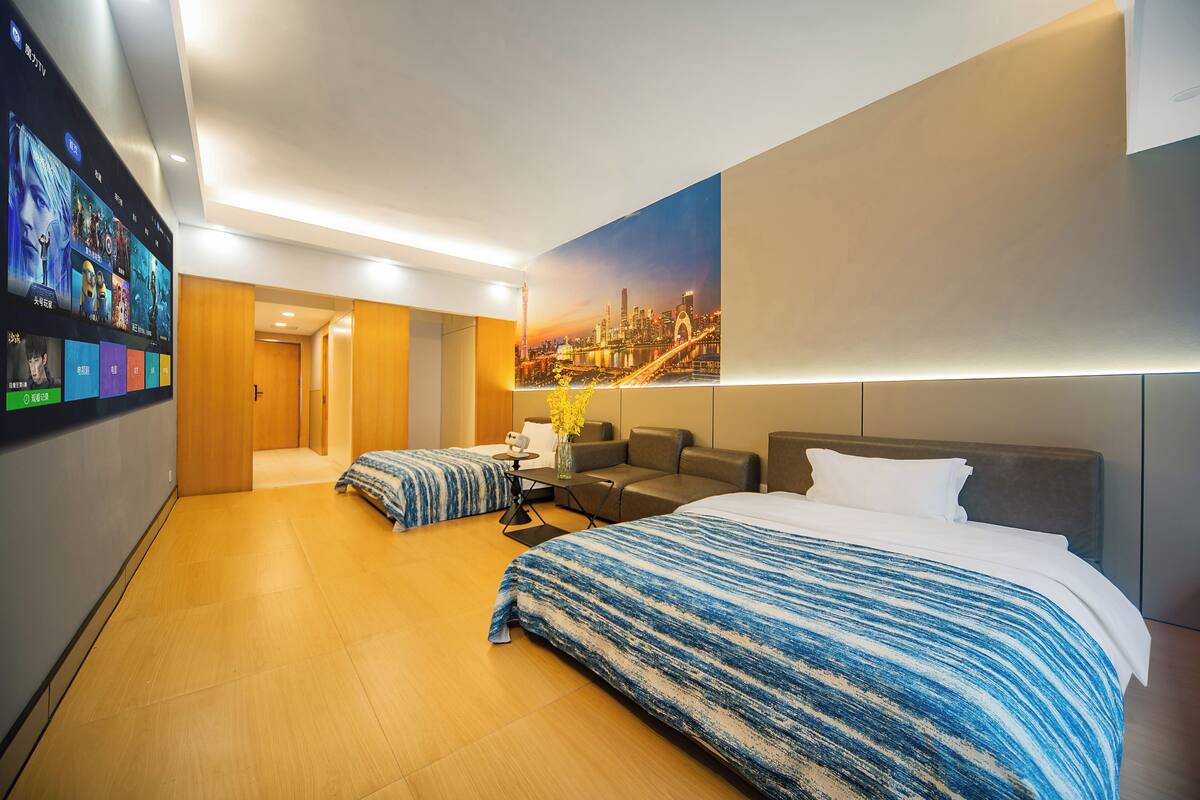 Habitación de hotel con una cama grande, un sofá, una mesita con un florero y un televisor de pared que muestra opciones de películas.