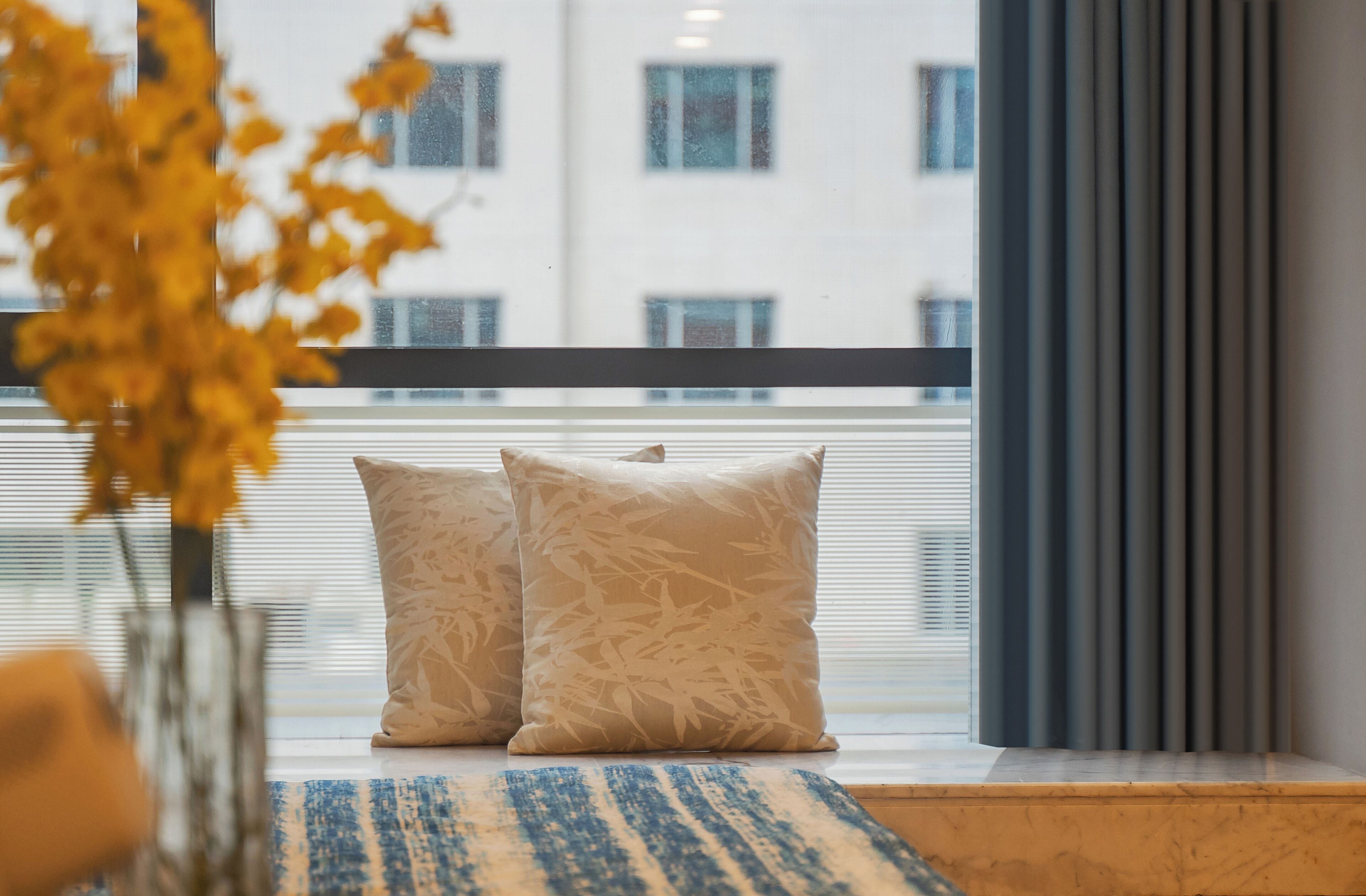 A room with a large window, two patterned pillows on a blue and white striped rug, and a vase with yellow flowers.
