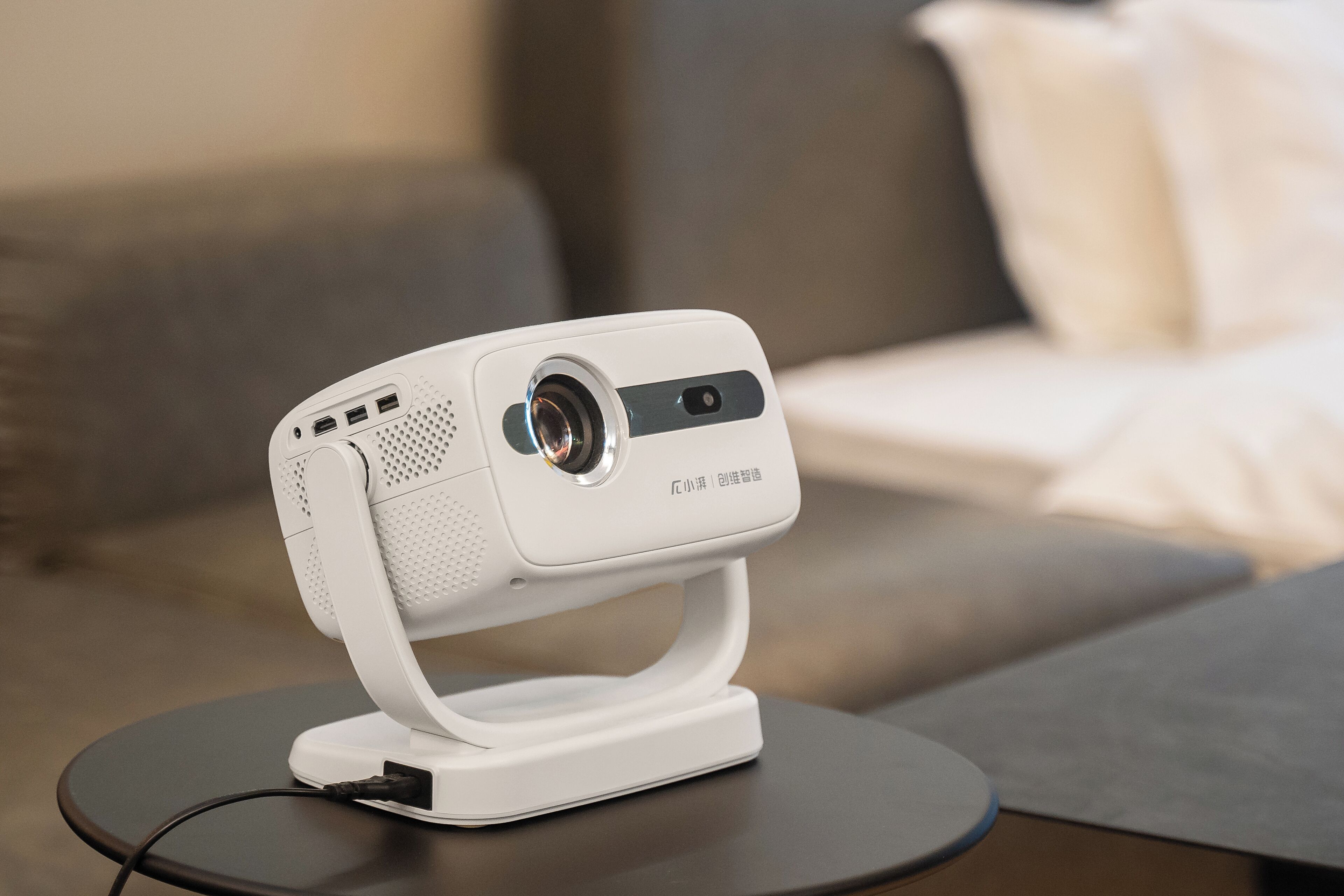 A white projector on a small table in a living room with a sofa and a cushion in the background.